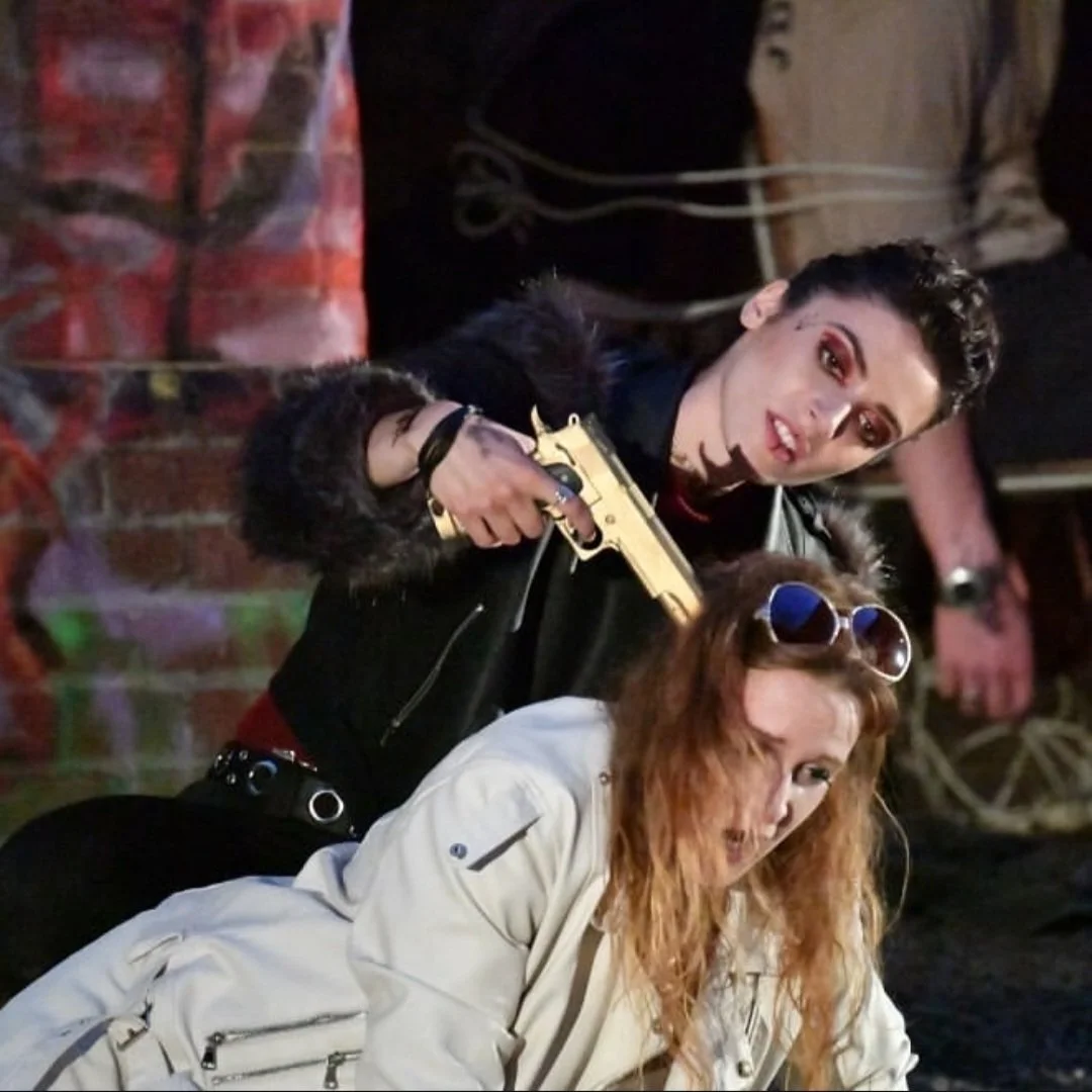 A woman with red hair wearing sunglasses and a beige jacket, and a woman with dark hair wearing dark makeup and a black leather jacket, holding a gold toy gun in a dramatic scene with a brick wall background.