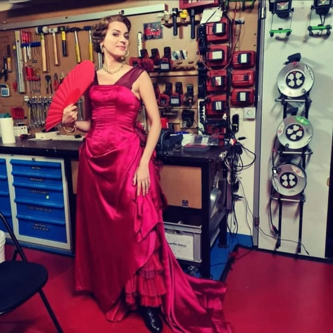 A woman in a vibrant pink gown poses in a workshop with tools and equipment on the wall behind her.