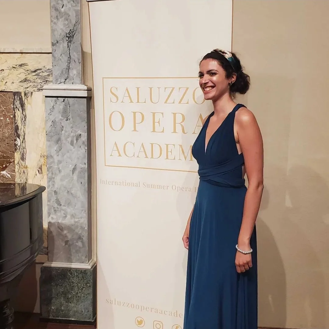 A woman in a sleeveless blue gown standing next to a sign that reads 'Saluzzo Opera Academy' at an event.