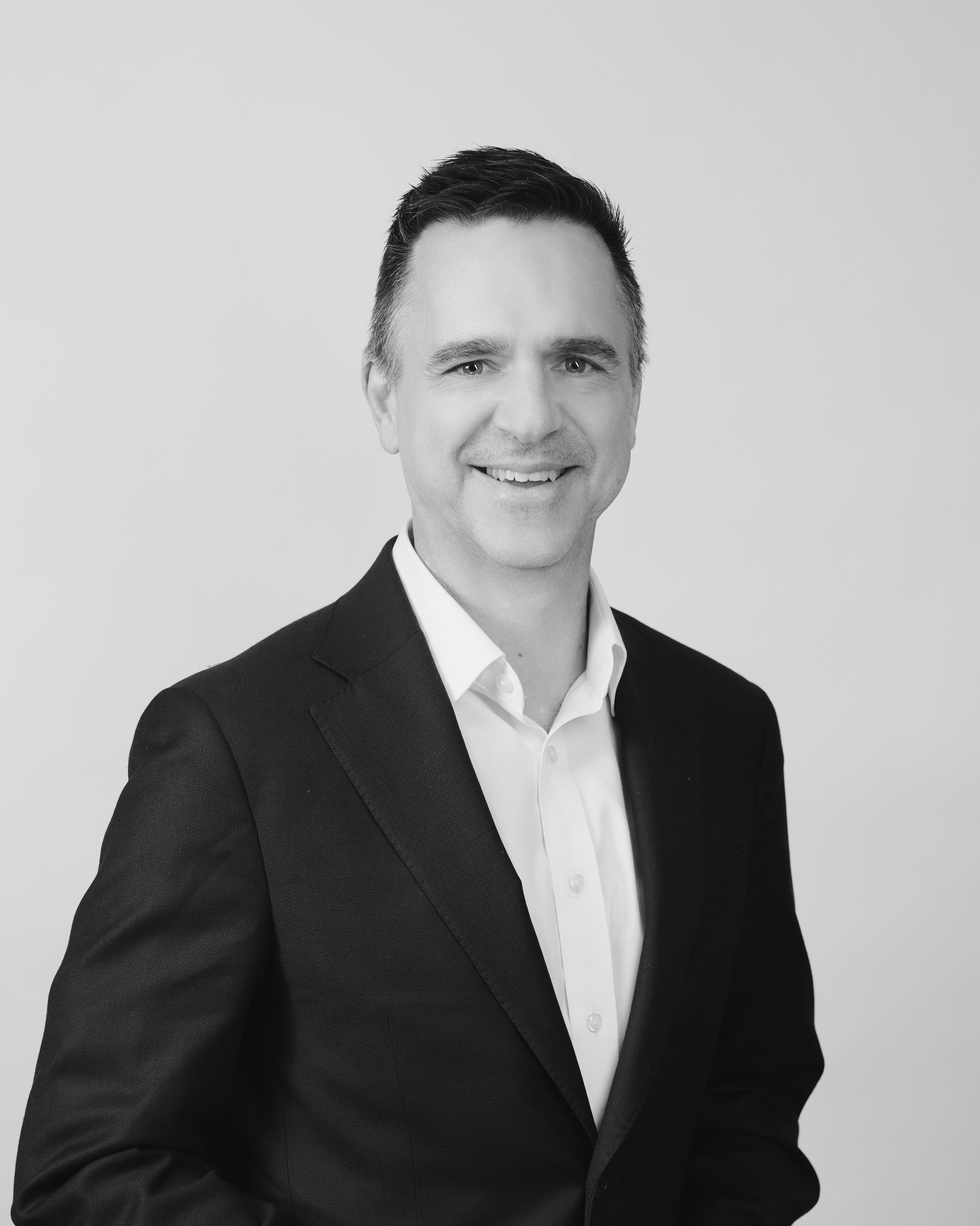 Jon Black, Partner at Oreana Advice Partners Black in a dark suit and white shirt, smiling against a plain light background.