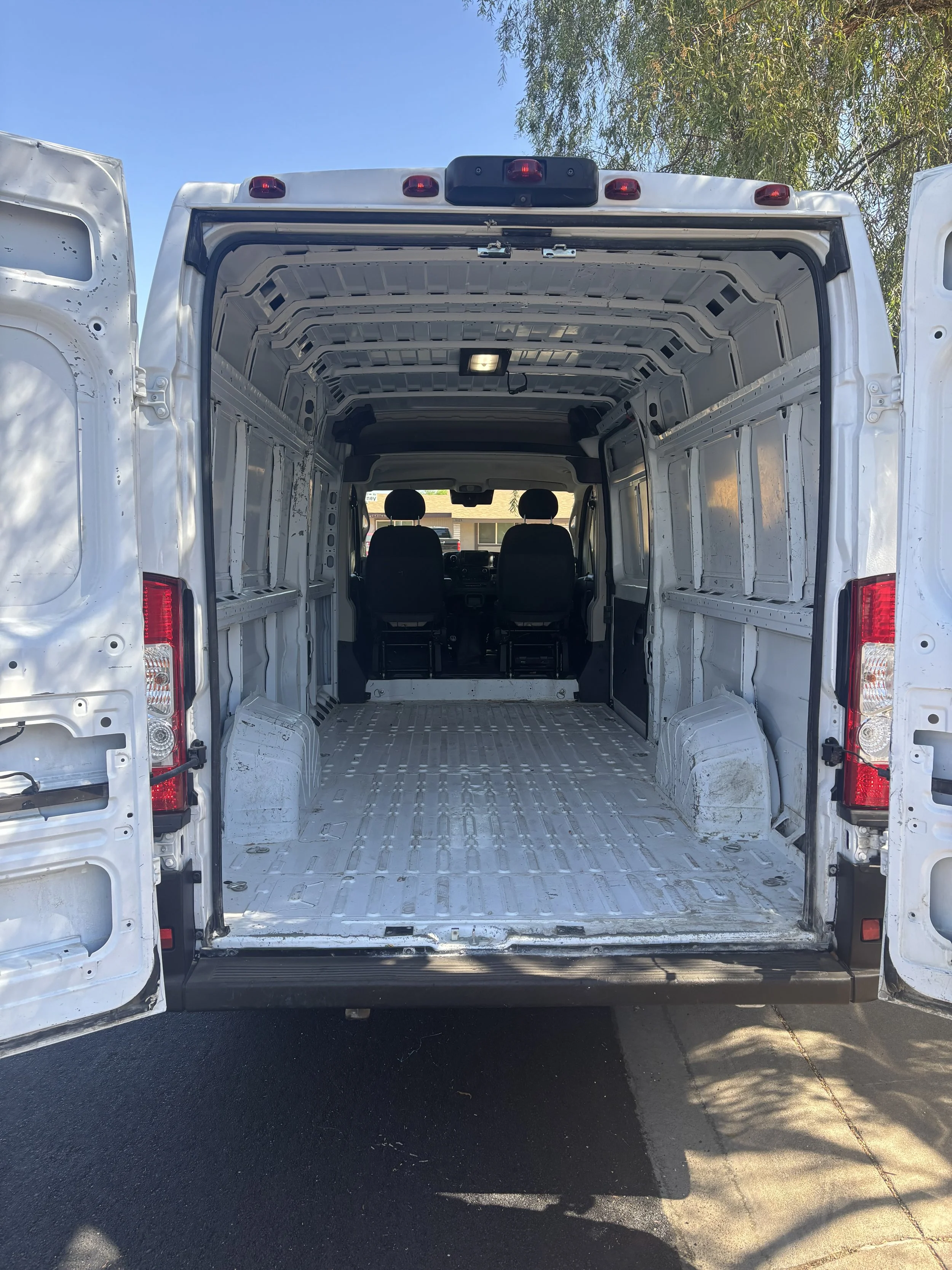 The image shows the open back of a white cargo van, with the interior empty and stripped down, sitting on a paved surface with a tree and a building visible in the background.