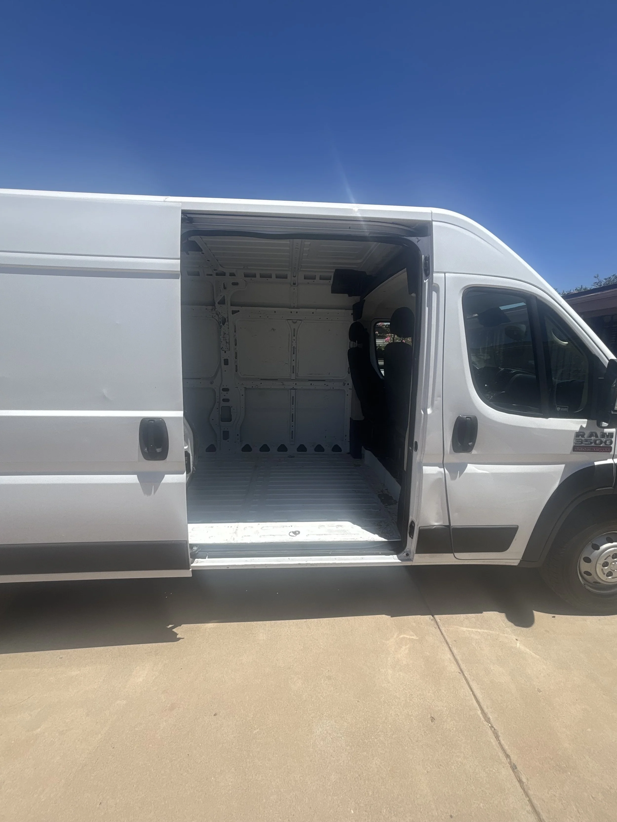 White cargo van with an open side door showing an empty cargo area, parked outdoors on a concrete surface under a blue sky.