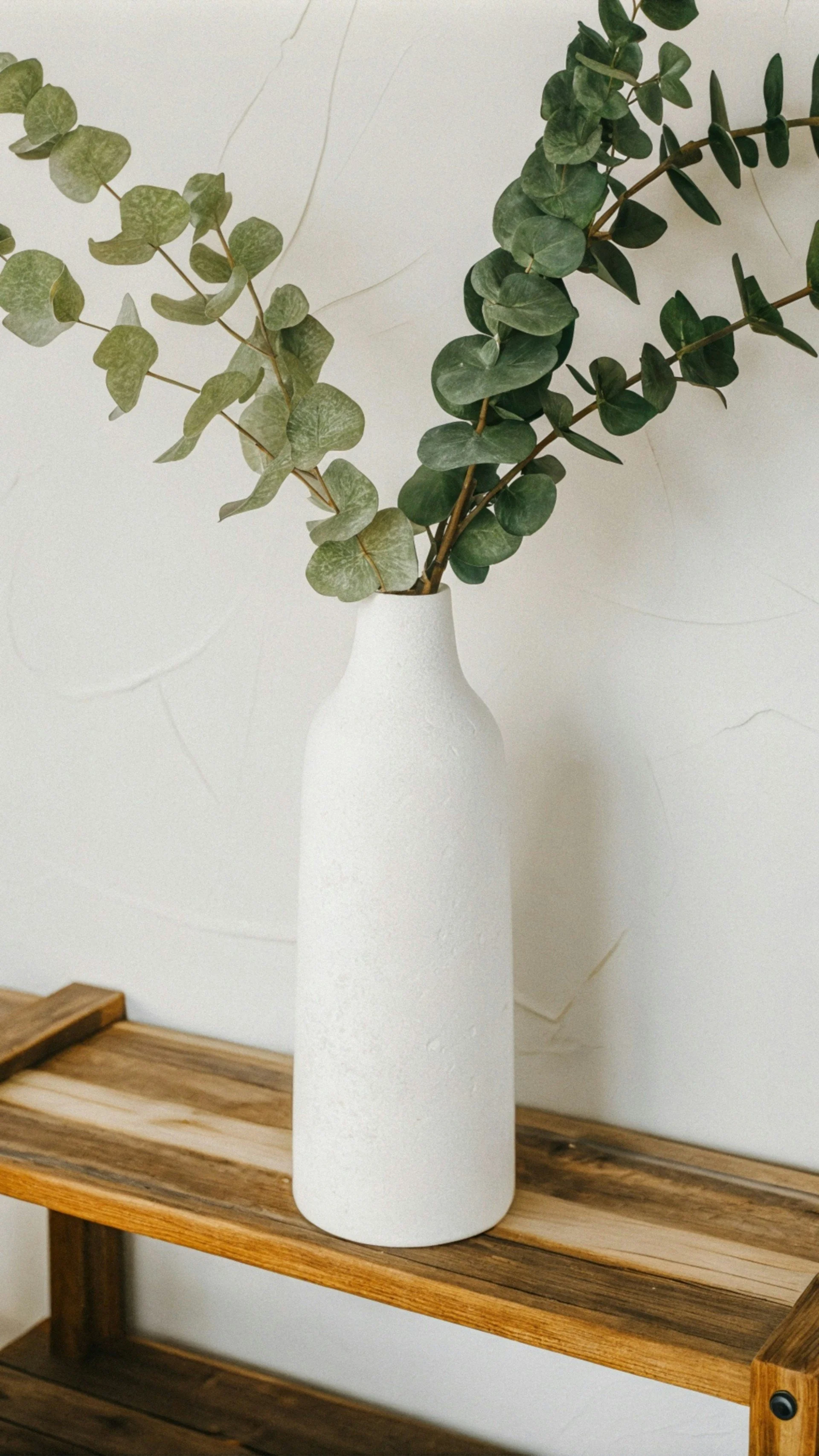 A white ceramic vase with a rough texture holding green eucalyptus branches, placed on a wooden shelf against a white wall.