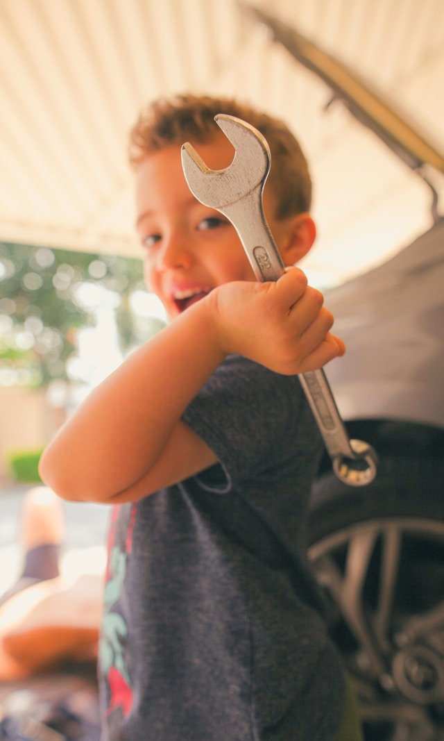 A young boy smiling and holding a large wrench in front of a vehicle.