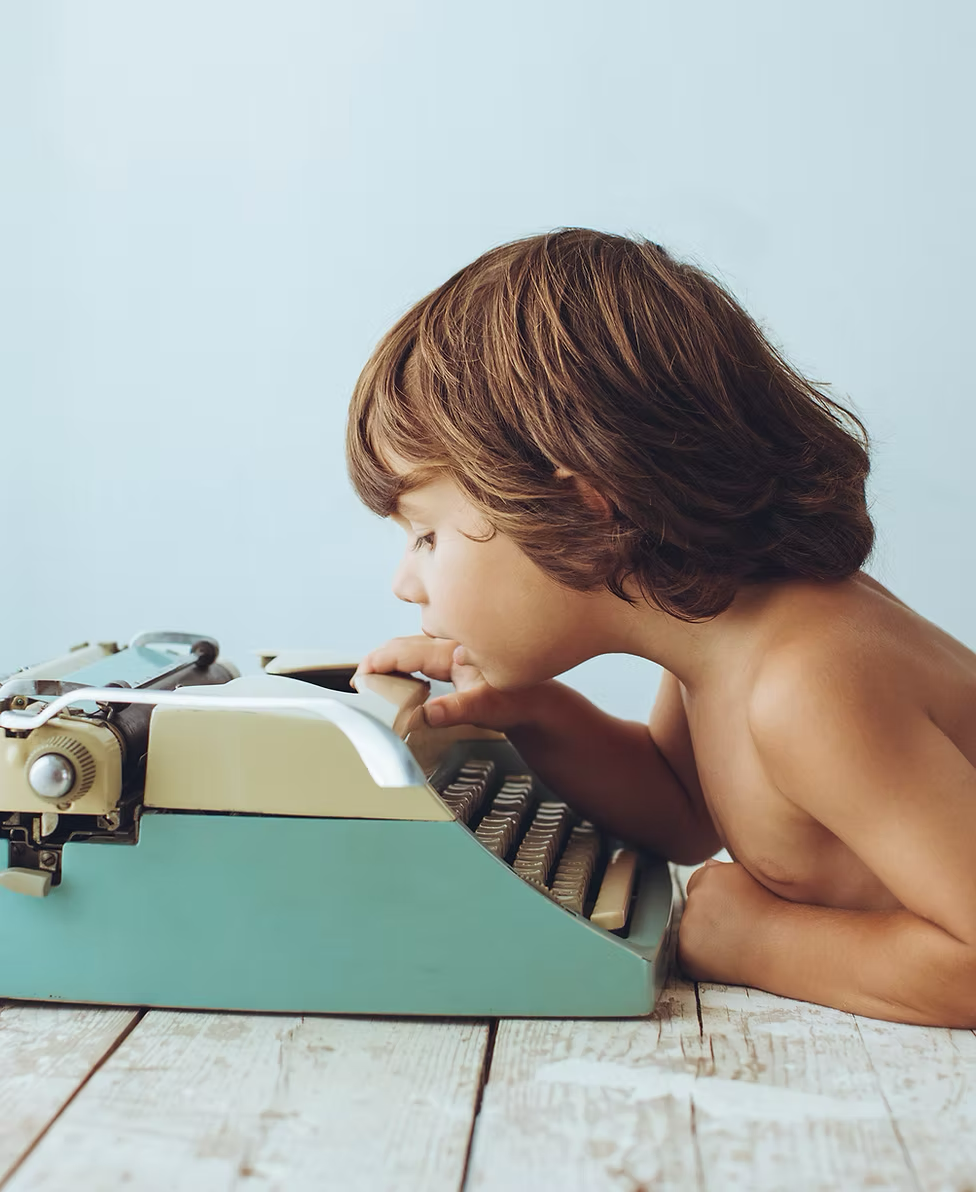 A boy with short brown hair is leaning over a vintage teal typewriter, appearing to be typing with his face very close to the keys.