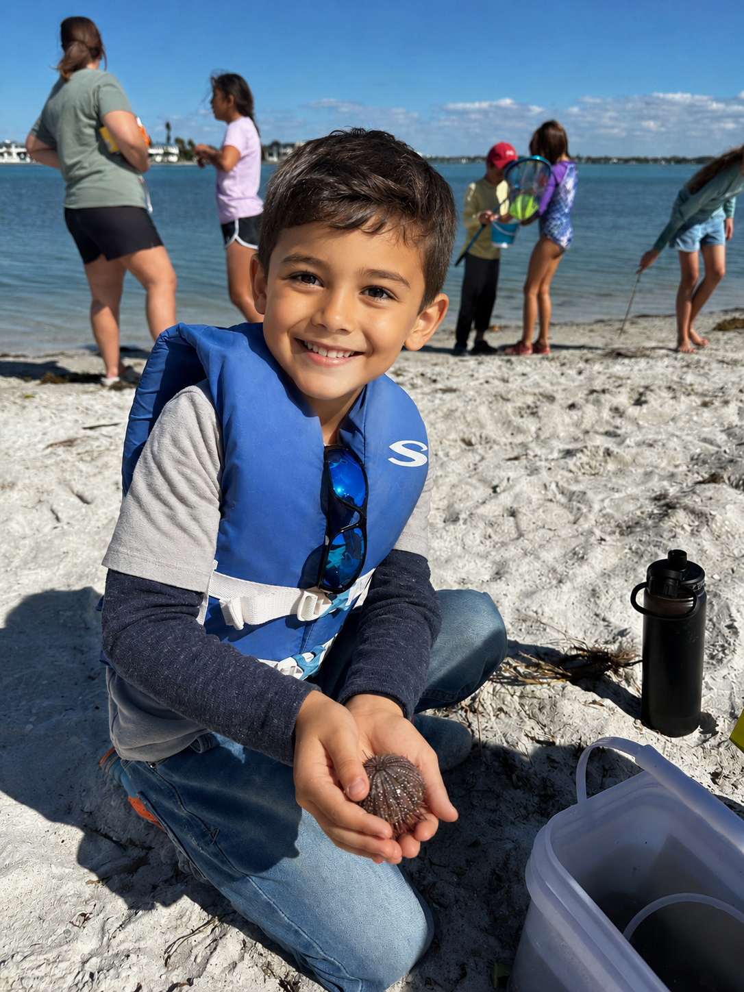 Smiling boy wearing a blue life jacket on the beach, holding a sea urchin, with children and water in the background.