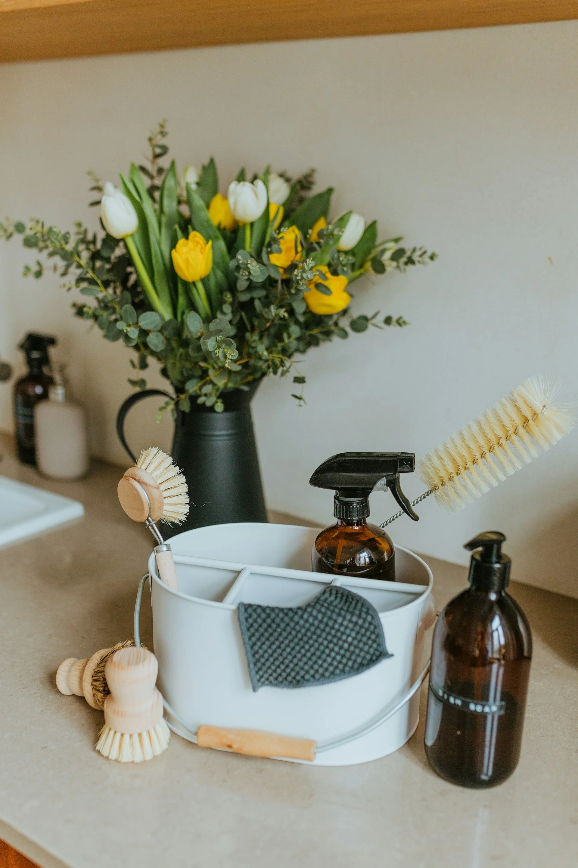 A kitchen countertop with a white basket holding cleaning brushes, a gray cloth, and amber spray bottles, with a background of a flower vase with yellow and white tulips and greenery.