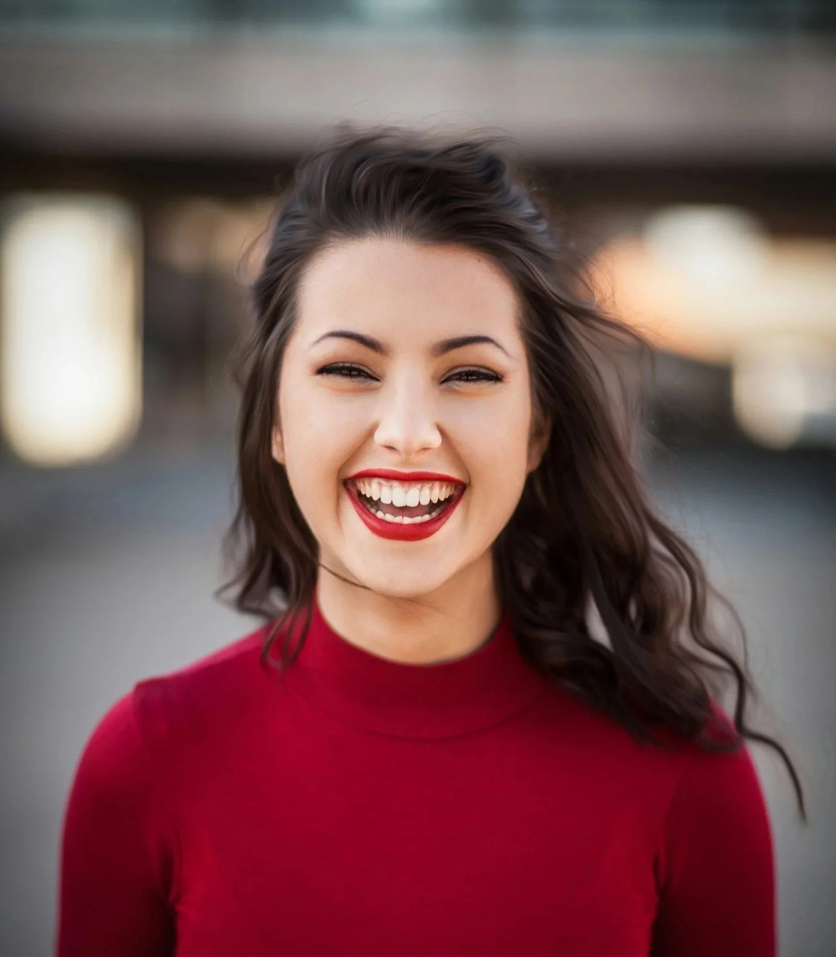 Close-up of a smiling woman with dark hair, wearing a red top, in an outdoor setting.