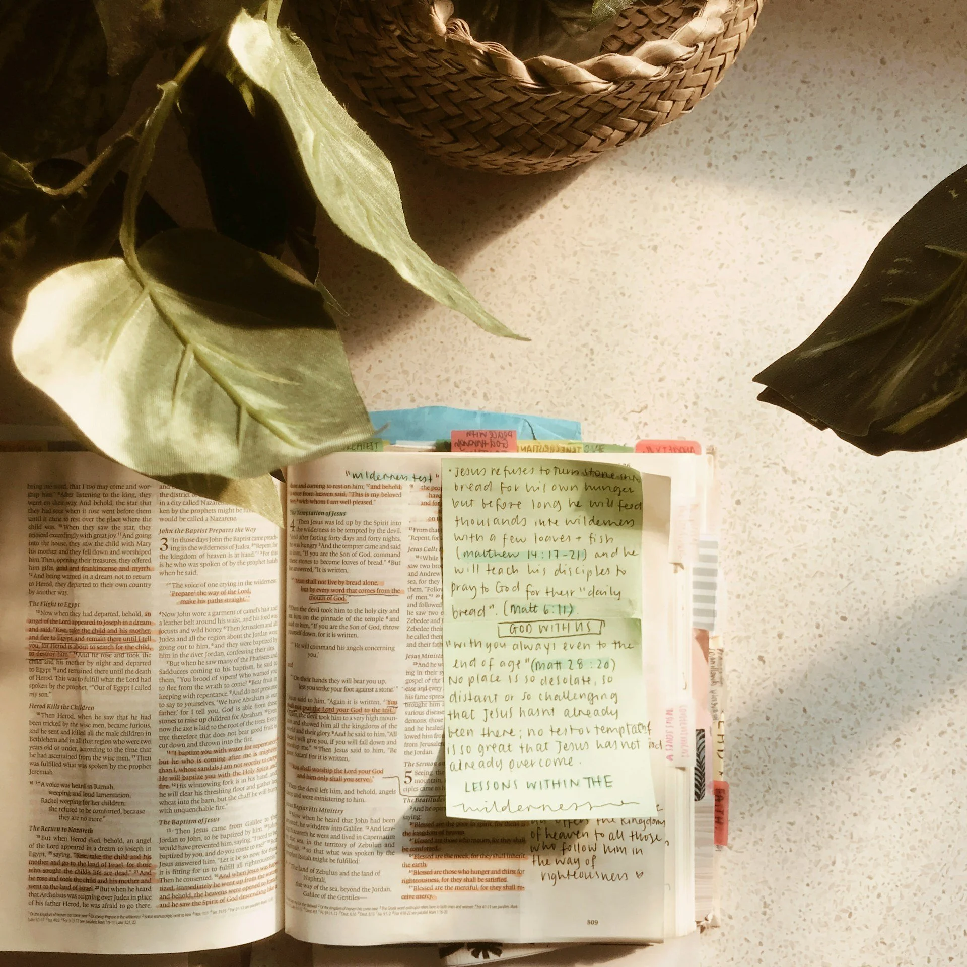 Open Bible with handwritten notes and highlighted scripture verses, decorated with green leaves, on a speckled countertop with a woven basket and black shoes nearby.