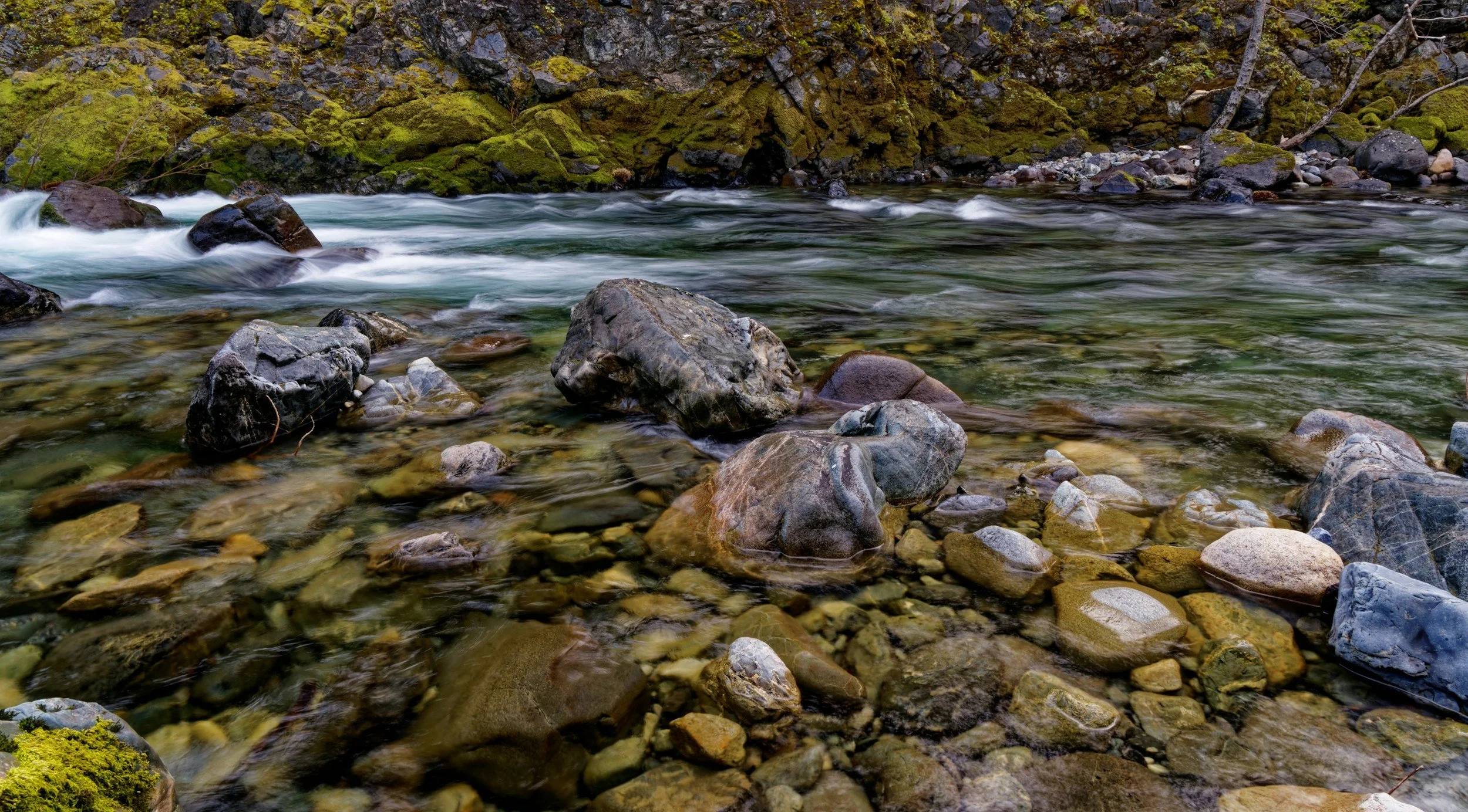 Flowing river with rocks and moss-covered hillside in the background