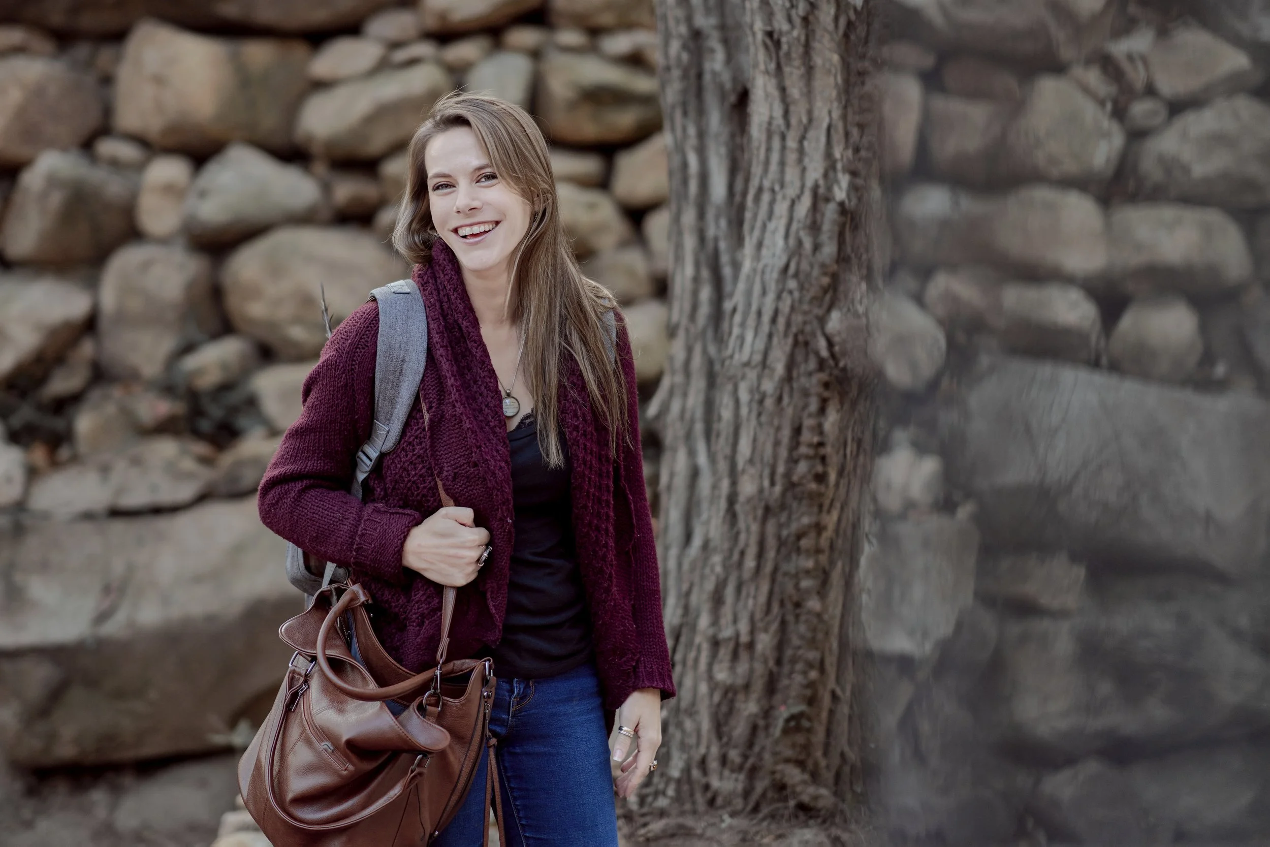 A woman with long light brown hair, wearing a burgundy cardigan, black top, and blue jeans, smiling while standing outdoors next to a tree with a stone wall in the background. She has a gray backpack over one shoulder and a large brown bag hanging from her arm.