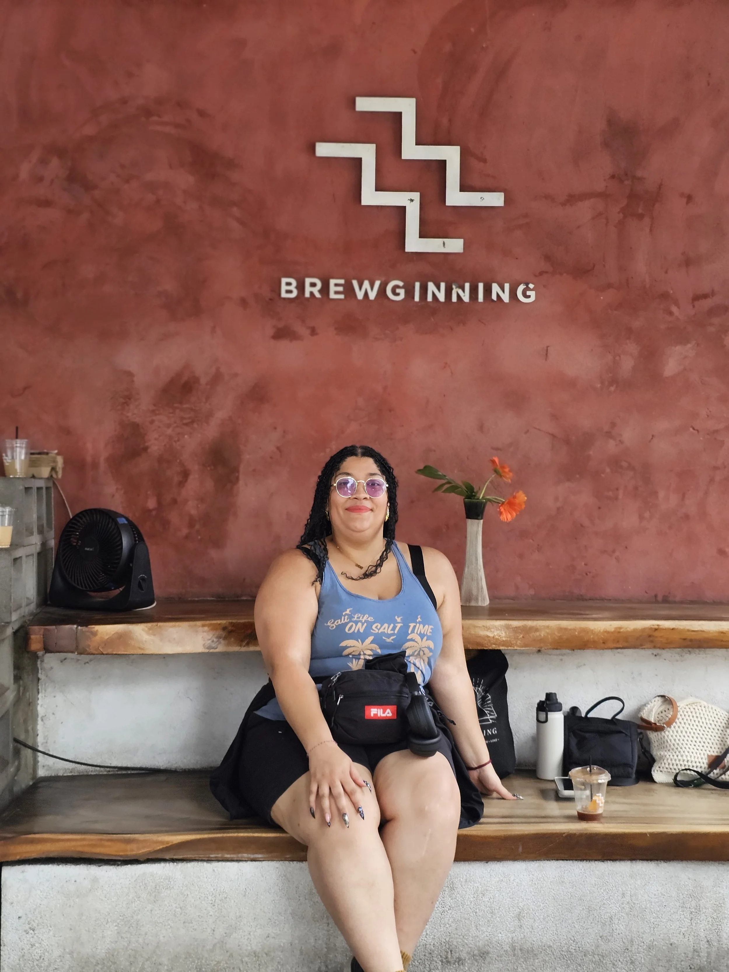 A woman with glasses and long dark hair sitting on a wooden bench in front of a red textured wall with the word 'BREWGINNING' and a minimalist mountain-like logo above it. She is wearing a blue tank top, black shorts, and has a black Fila bag on her lap. There is a vase with orange flowers on her right, a water bottle, and other personal items on the bench.