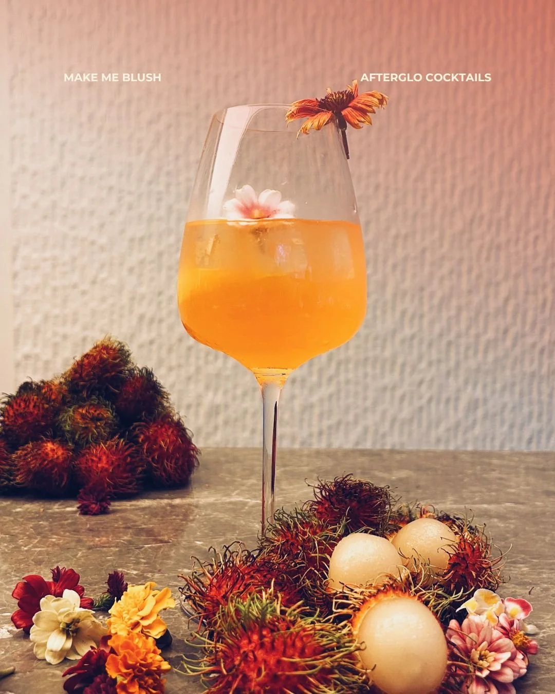 A cocktail glass filled with an orange drink, garnished with a small flower and a decorative straw, placed on a table surrounded by rambutans, dragon fruit, and various flowers.