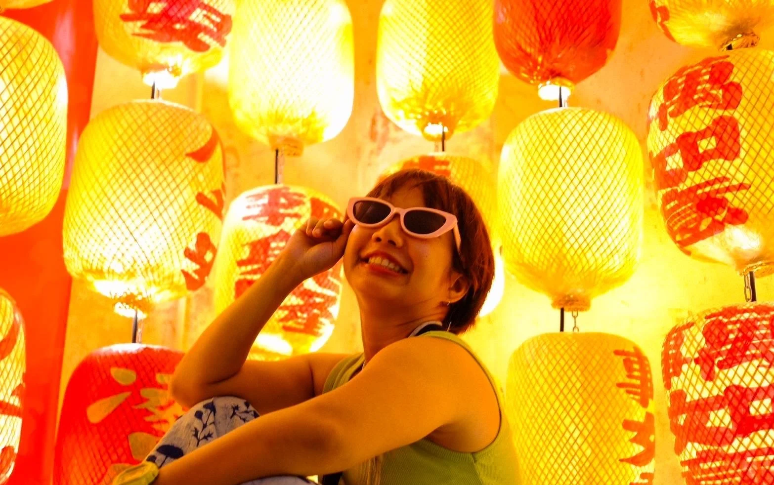 A smiling person wearing sunglasses, seated in front of a wall decorated with illuminated yellow and red paper lanterns.