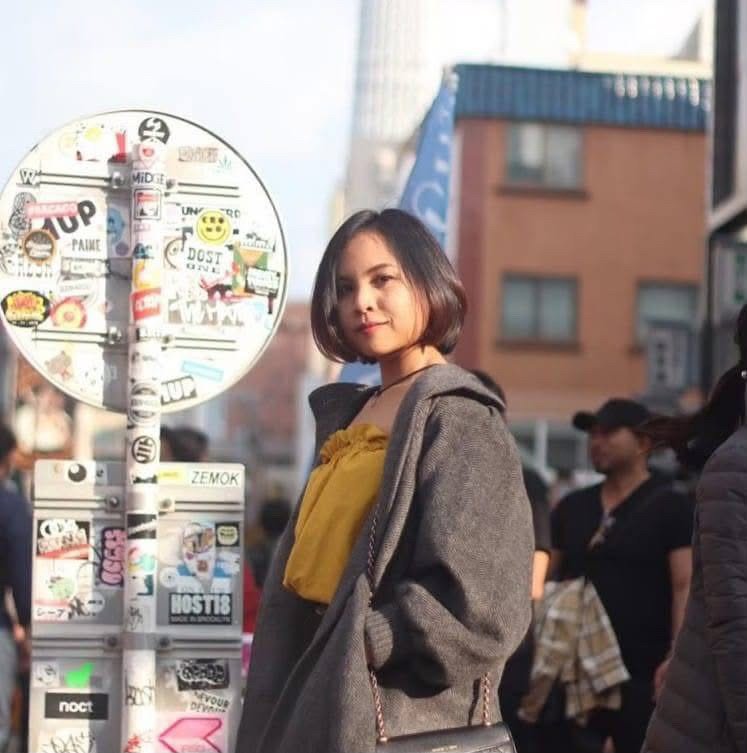 A young woman with short dark hair wearing a yellow top and gray coat stands on a city street near a sticker-covered pole, with buildings and pedestrians in the background.