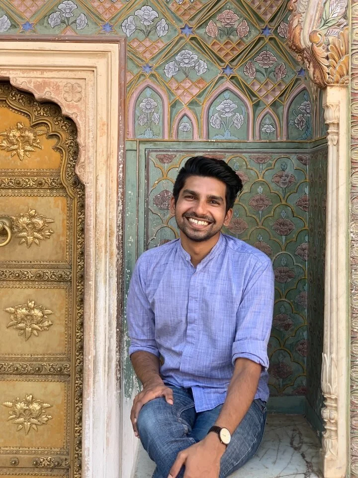 A smiling man with dark hair and a beard sitting on a marble surface in front of a decorated wall with floral patterns and intricate carvings.