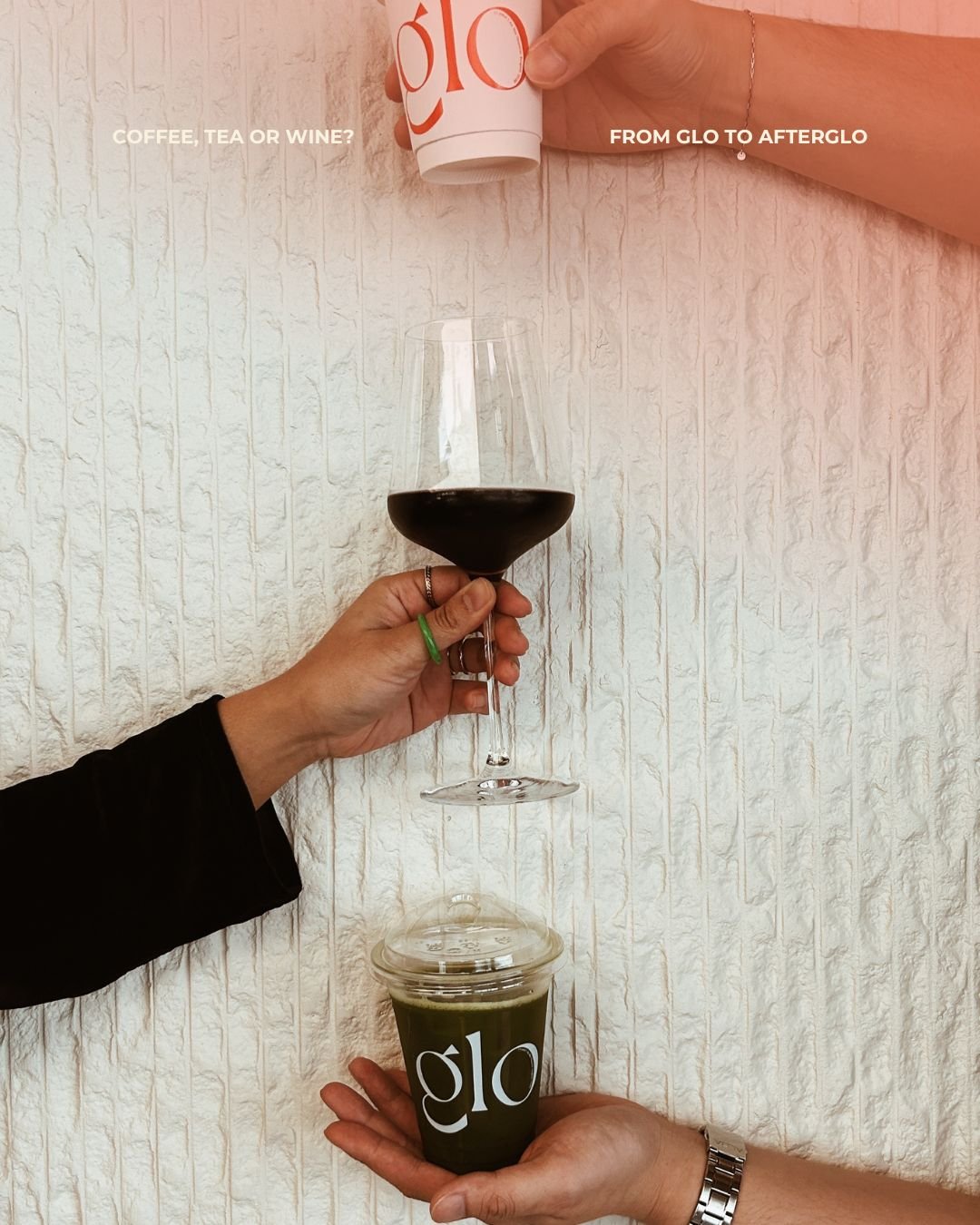 A layered drink setup with a coffee or matcha in a plastic cup labeled 'glo' at the bottom, topped with a clear dome lid. Above, a person holds a glass of red wine, and another person's hand is pouring a drink from a cup labeled 'glo' into the wine glass. The background is a textured white wall.