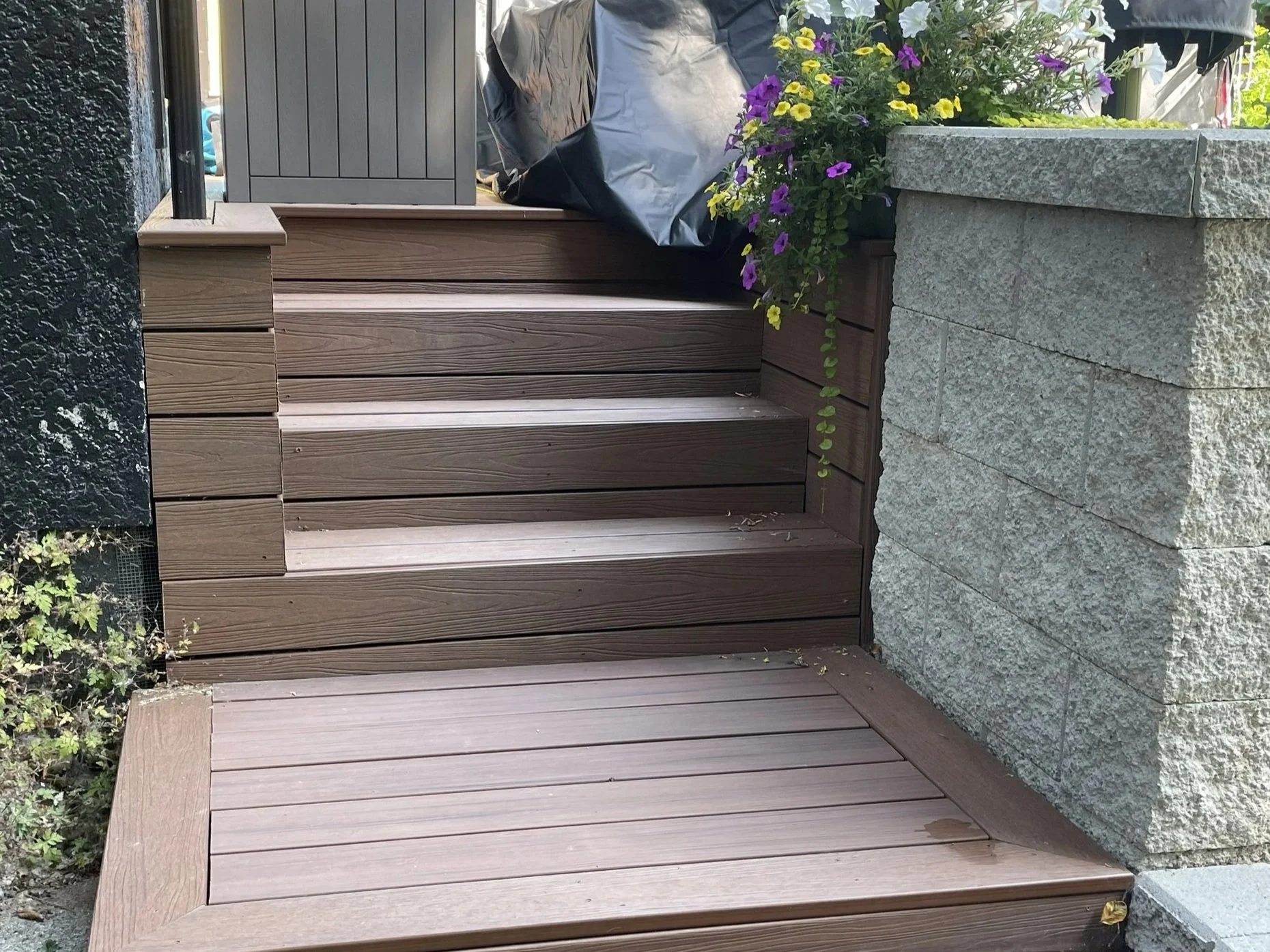 A wooden fence with horizontal slats, a stone planter with colorful flowers, and a stone or concrete tile path.