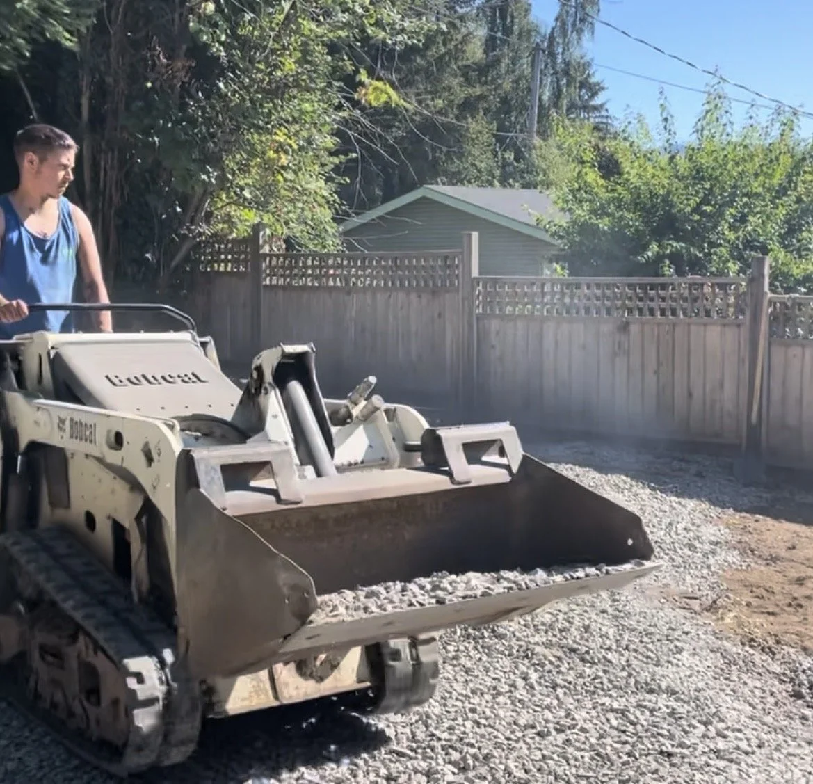 A man operating a Bobcat mini excavator with gravel in the bucket, working on a construction project in a backyard enclosed by a wooden fence, with trees and a house visible in the background.