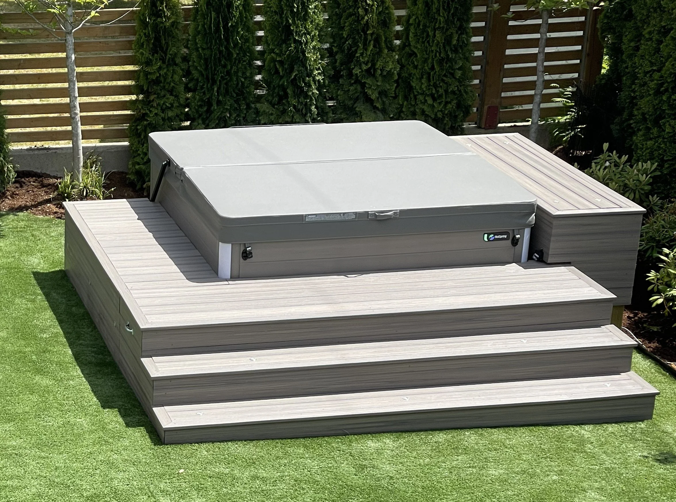 A backyard with a large rectangular hot tub covered with a gray cover, placed on a light-colored wooden deck. The deck has steps leading up to it. Surrounding the area are green grass, various plants, and tall, evenly spaced evergreen trees. A wooden fence encloses the backyard.