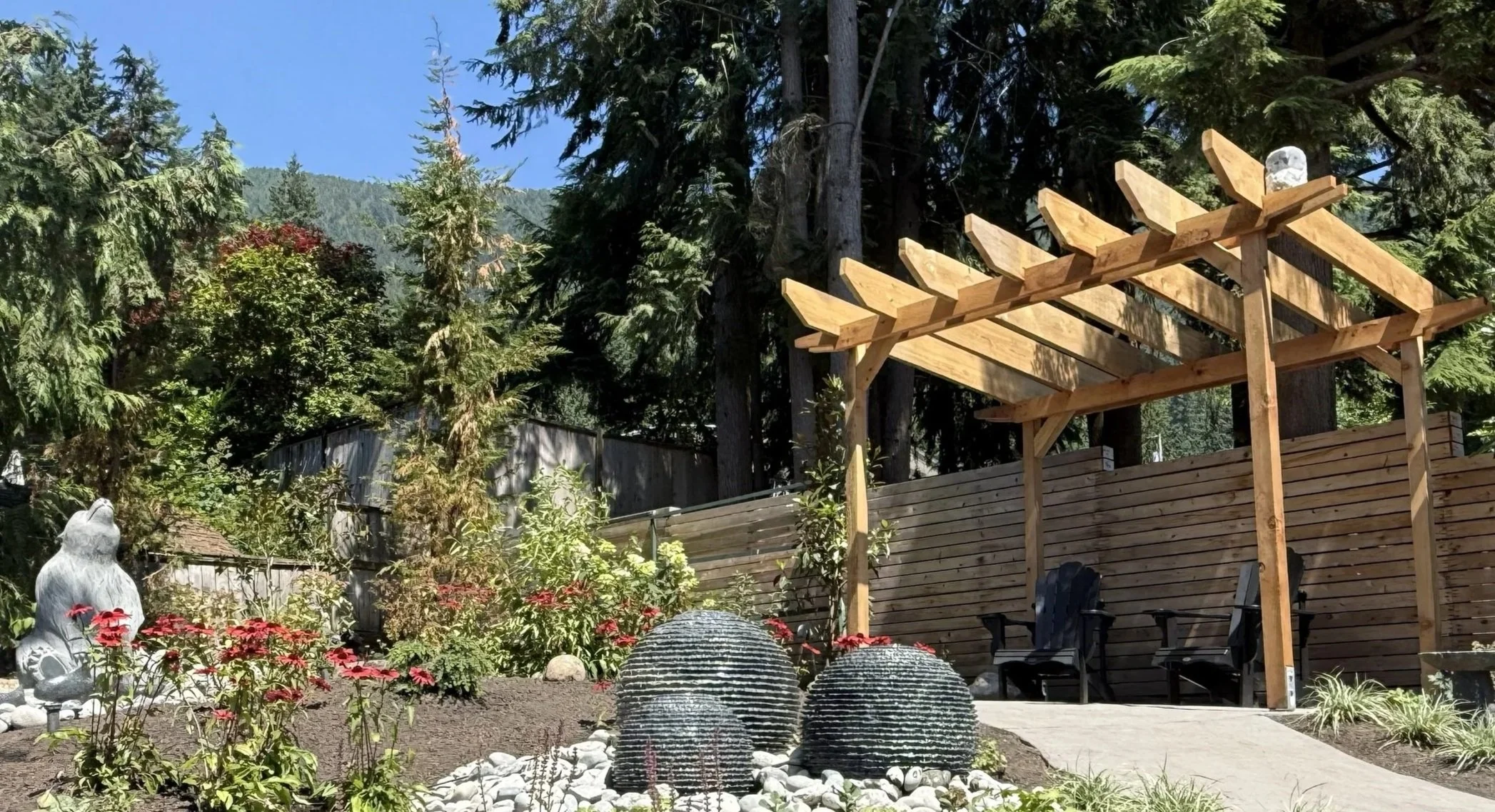 A backyard garden with decorative stone fountains, red flowers, and a wooden pergola under tall pine trees, with a mountain in the background.
