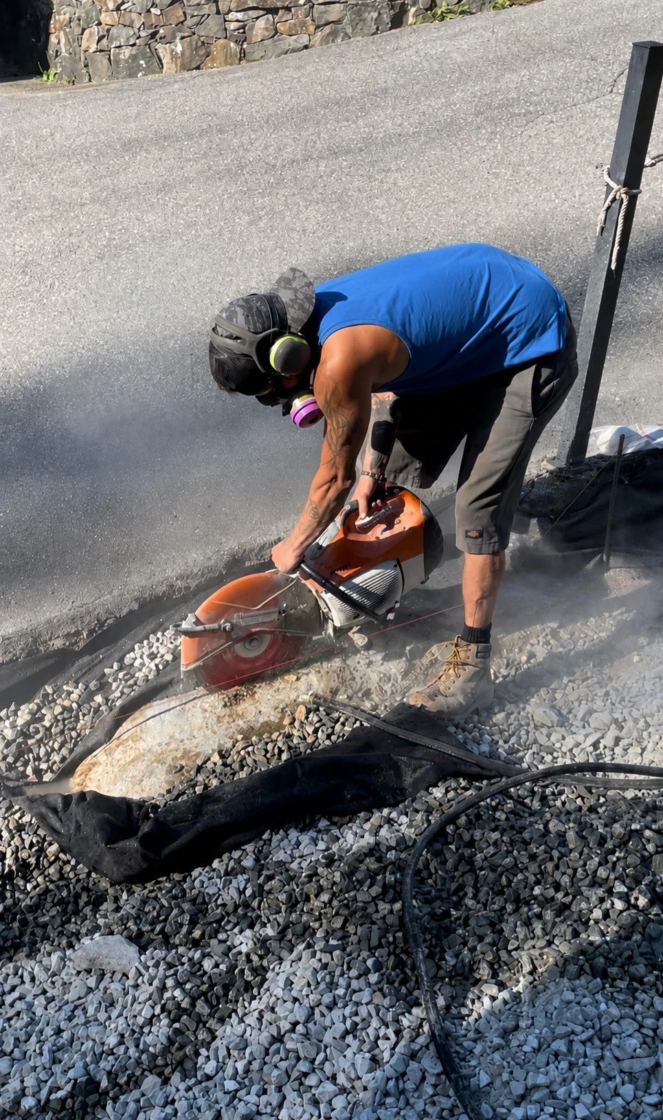 A person using a concrete cutting saw to cut a large stone on a gravel surface, wearing protective gear including a helmet with hearing protection and a mask.