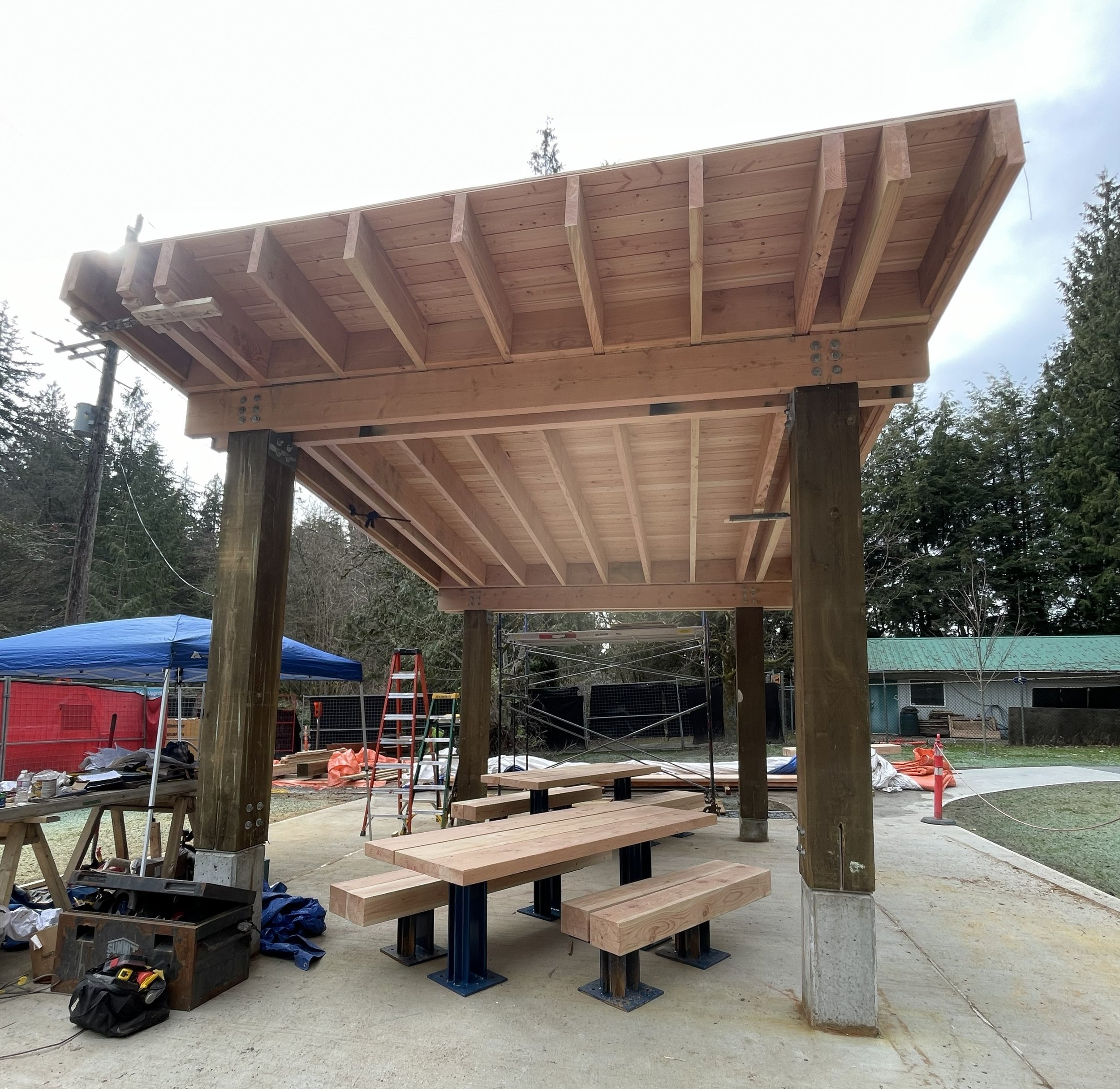 Under construction pavllion with support beams and tools at a construction site in a wooded area.