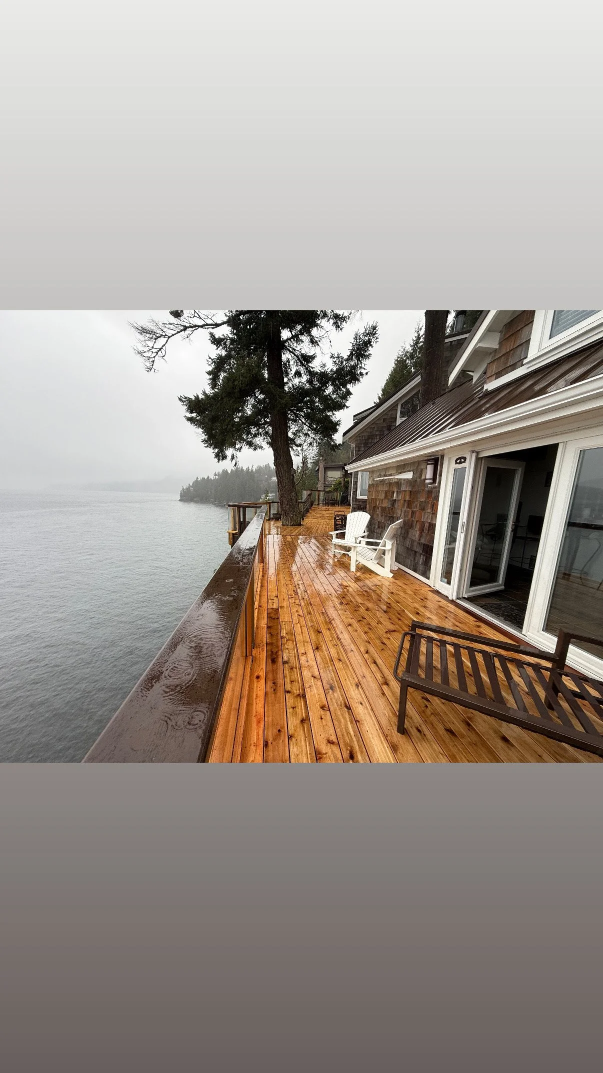 Wooden deck overlooking a body of water with trees in the background, and a house with large windows and sliding doors.