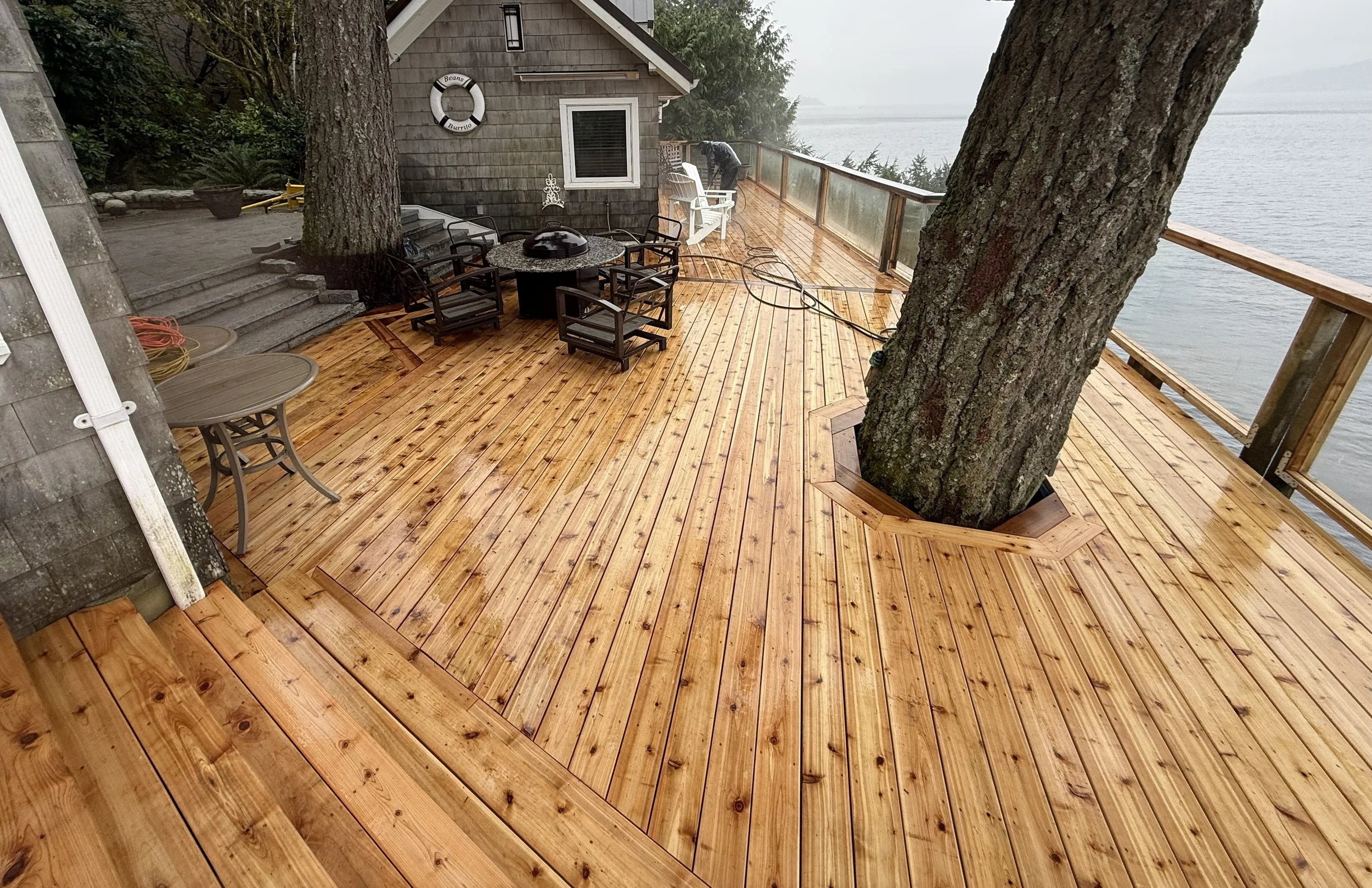 A wooden deck by the water with two large trees growing through it, outdoor furniture including a round table with a barbecue on top, chairs, and a white Adirondack chair. Overcast sky and foggy lake in the background.