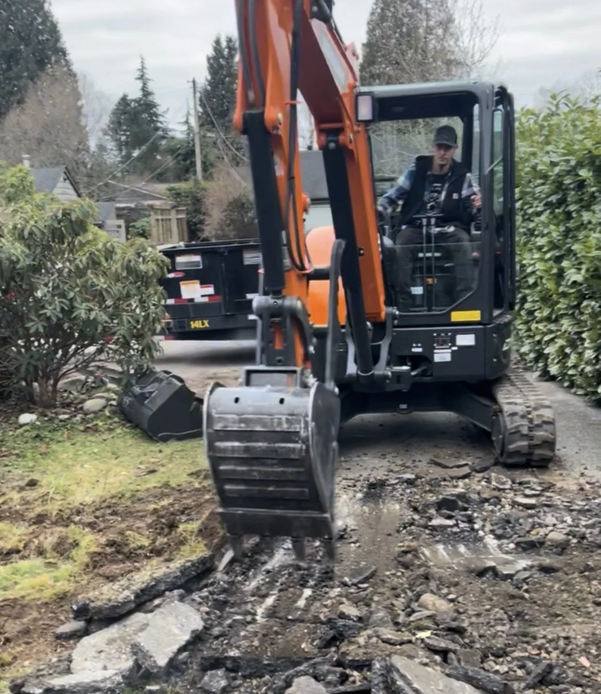 A person operating an orange and black mini excavator on a dirt path, breaking rocks near bushes in a residential area.