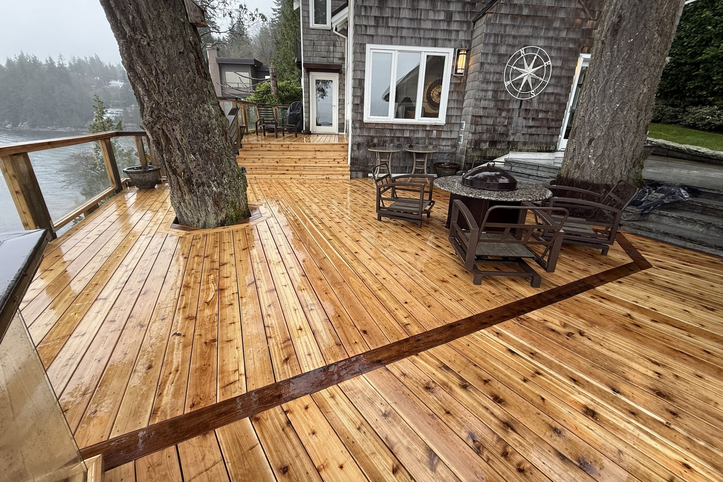 A wooden deck with trees growing through cutouts, outdoor furniture, and a view of water and distant houses in a foggy or rainy setting.