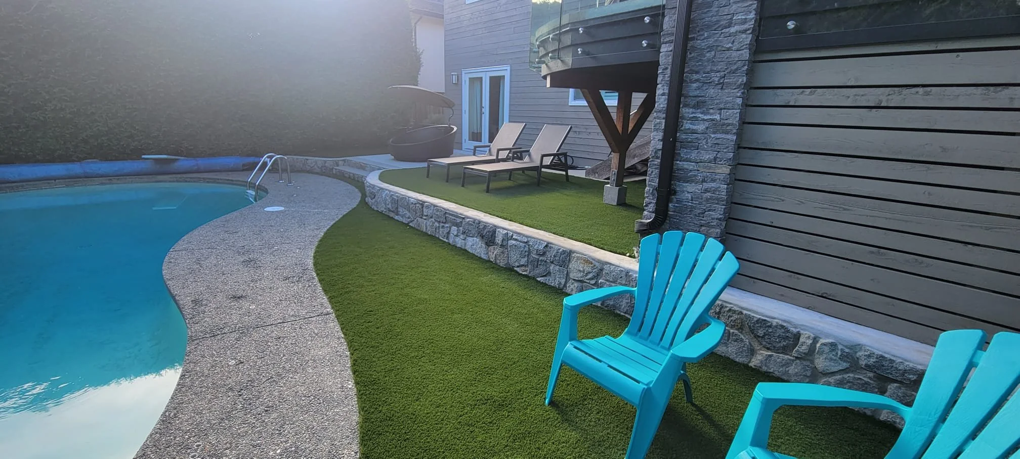 Backyard with a swimming pool, two lounge chairs, a blue Adirondack chair, artificial grass, and a gray wooden house wall.