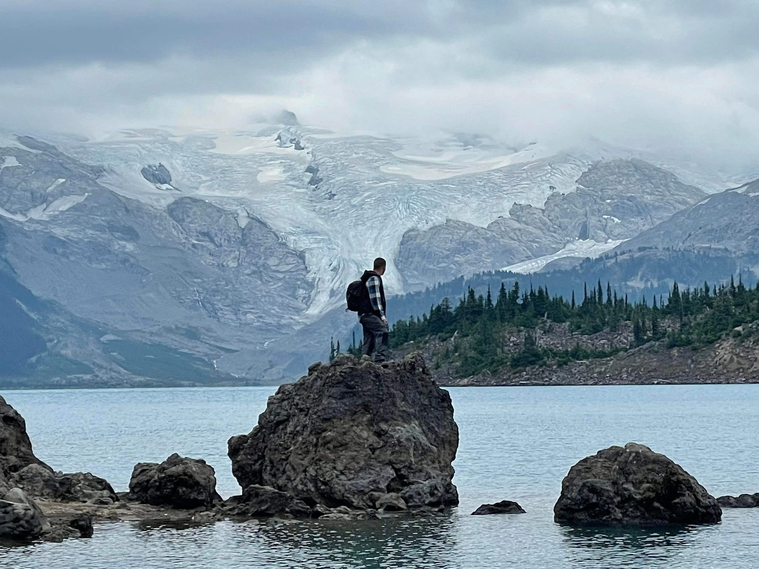 A person standing on rocks by a lake with mountainous, snow-covered peaks and cloudy skies in the background.