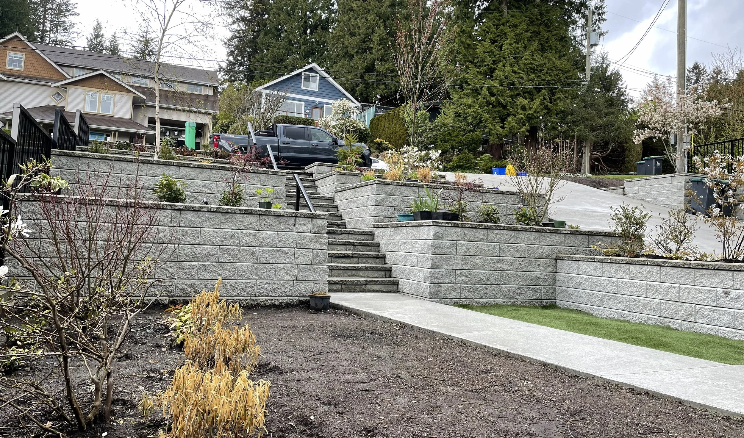 A residential hillside with concrete steps and retaining walls, a parked truck, and a house with blue siding surrounded by trees and shrubbery.