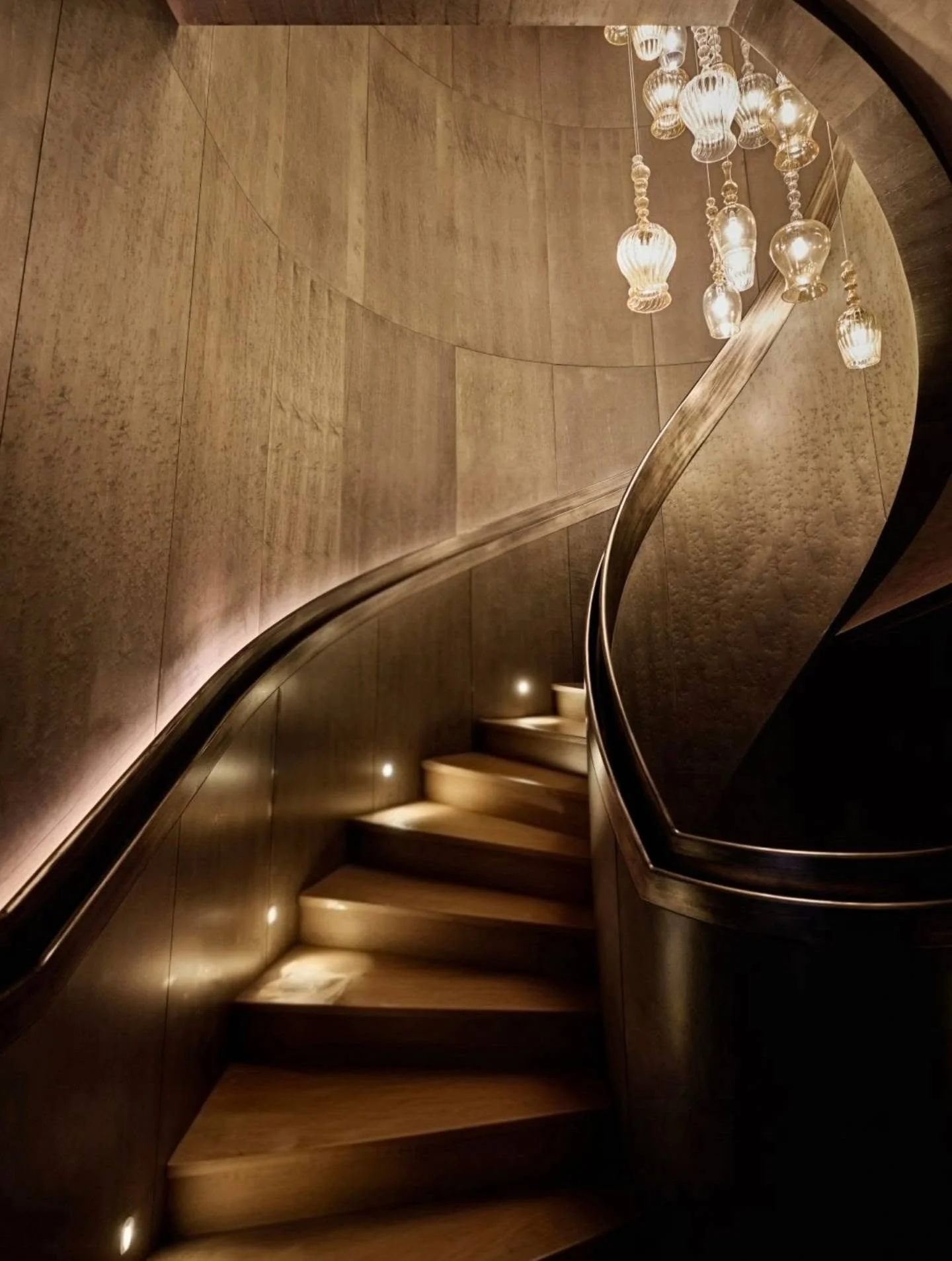 Rhythmic curves of a dark handrail and integrated lighting define a seamless helical stairway, where textured vertical panels create a dramatic backdrop under a shower of warm, hand-blown glass light fixtures.

​Interior Design by Tihany Design.

​Th