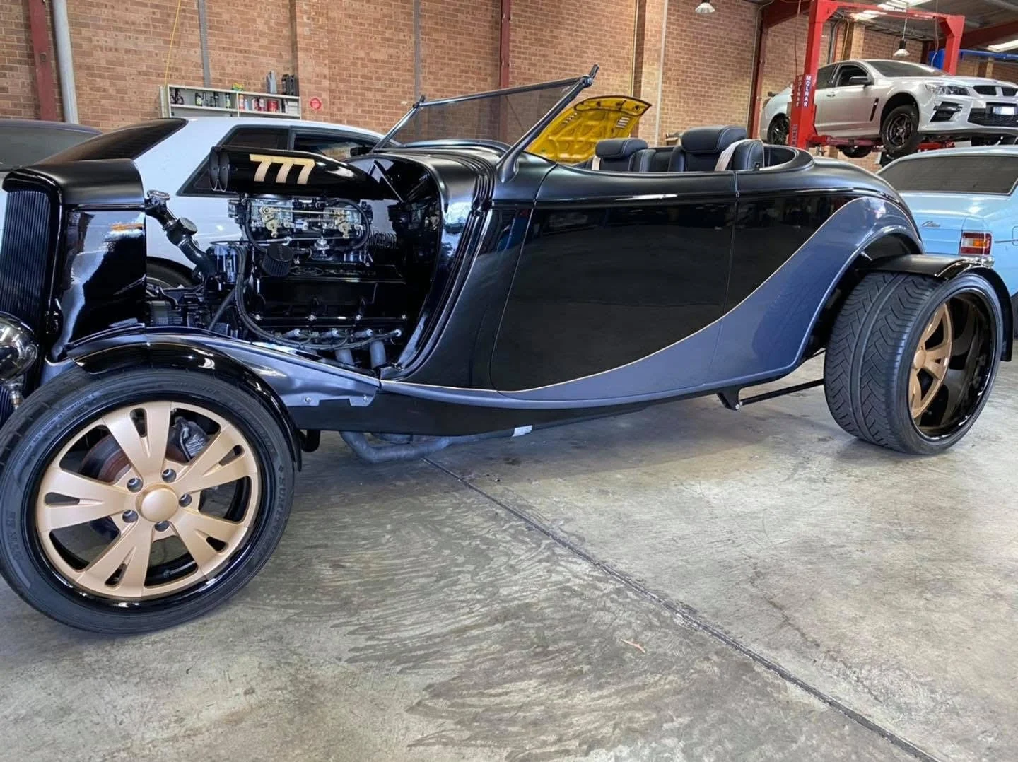 A black vintage convertible car with a supercharged engine on display in an indoor garage, surrounded by modern cars and automotive equipment.