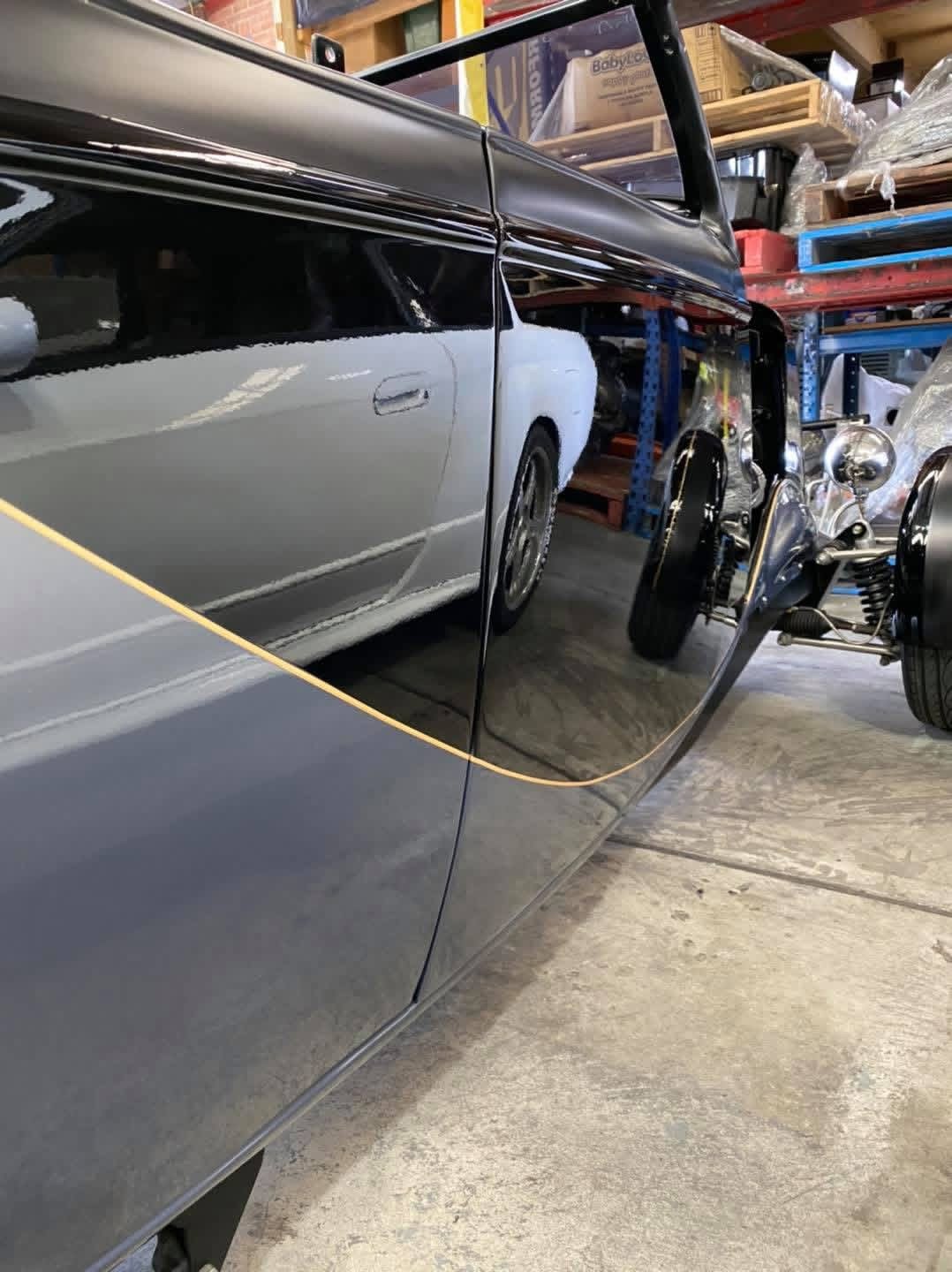 Reflected side view of a classic black and silver car in a garage, with tools and shelves in the background, alongside a vintage motorcycle.