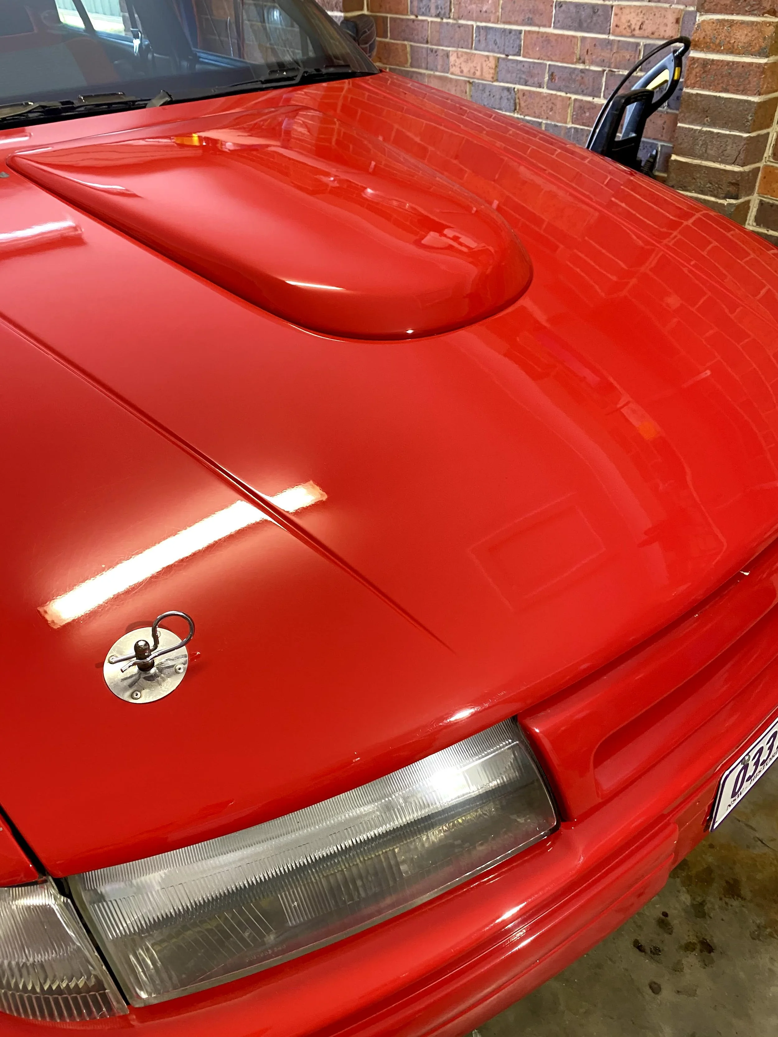 Close-up of a red car's front, showing the hood with a raised scoop, a side mirror, part of the grille, and a brick wall in the background.