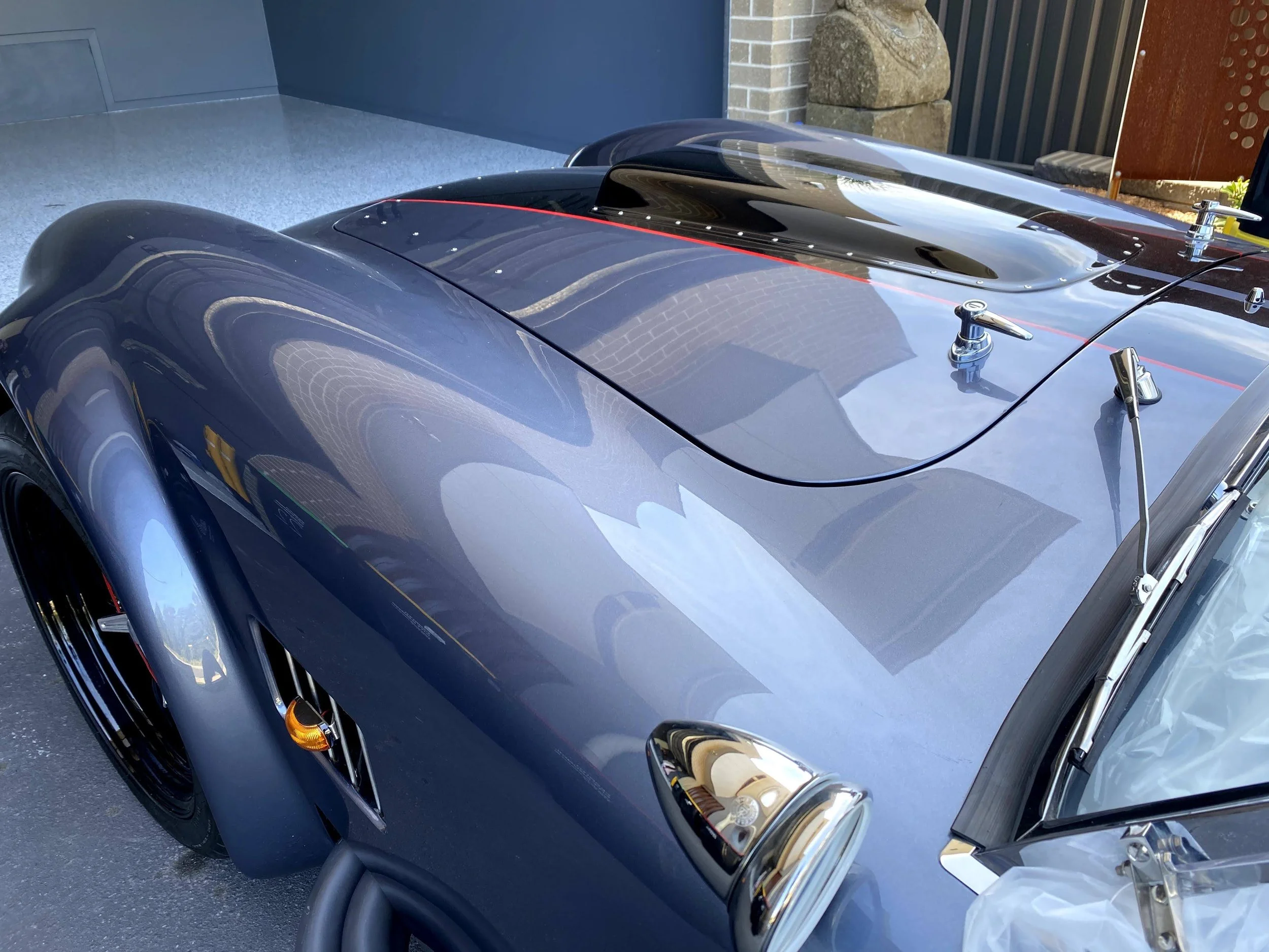 Close-up of a vintage convertible race car with a black body, chrome accents, and a red pinstripe, parked outdoors on a sunny day near a brick wall and sculpture.