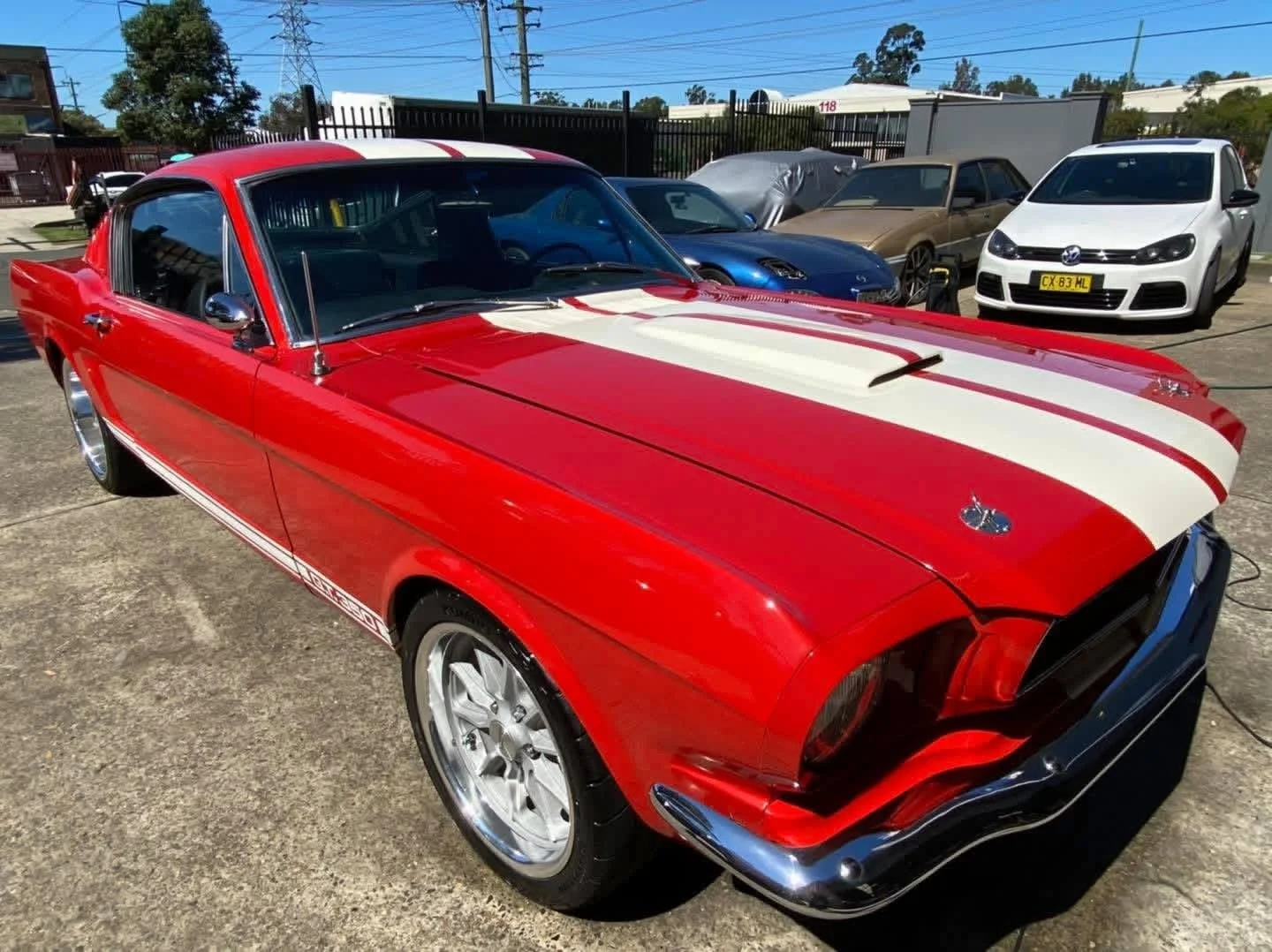 Red classic Ford Mustang with white racing stripes parked in a lot, surrounded by other cars. The car is a vintage model with shiny chrome accents and aftermarket wheels.