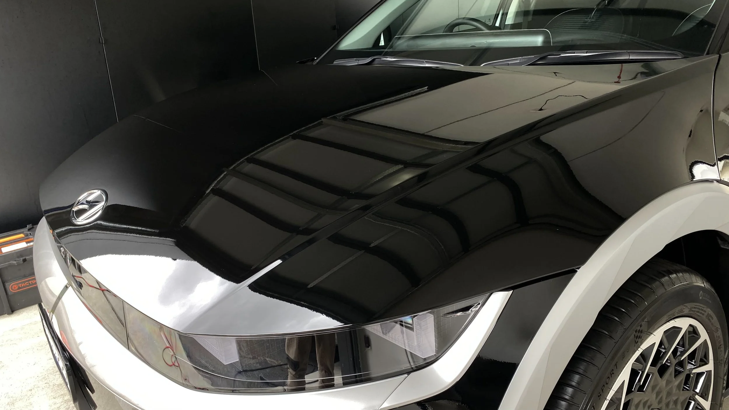 Close-up of the front part of a black and silver car, showing the hood, headlight, and part of the windshield and tire.