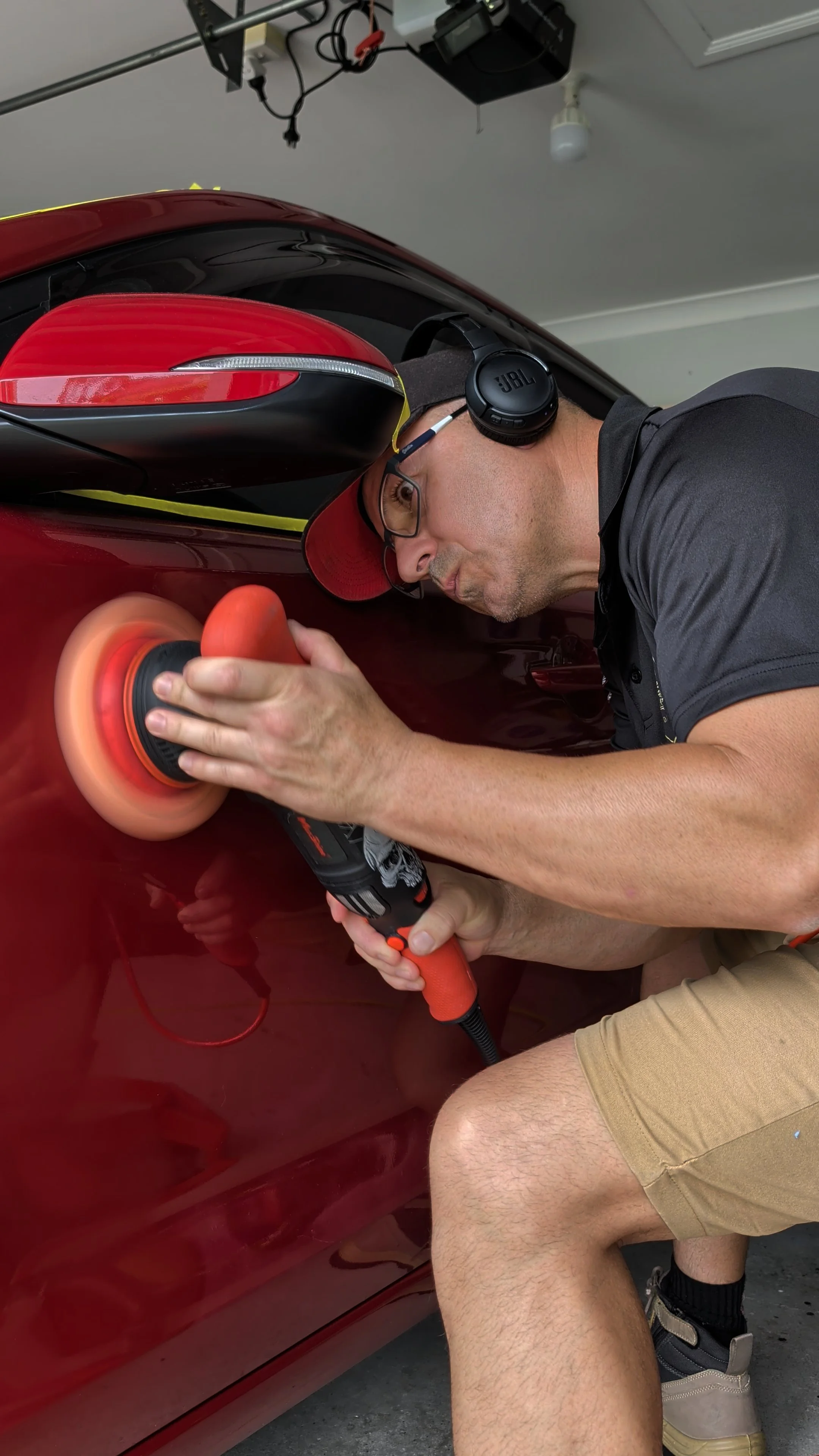 Man polishing a red car with an orange buffer pad in a garage, wearing glasses, a cap, and JBL headphones.