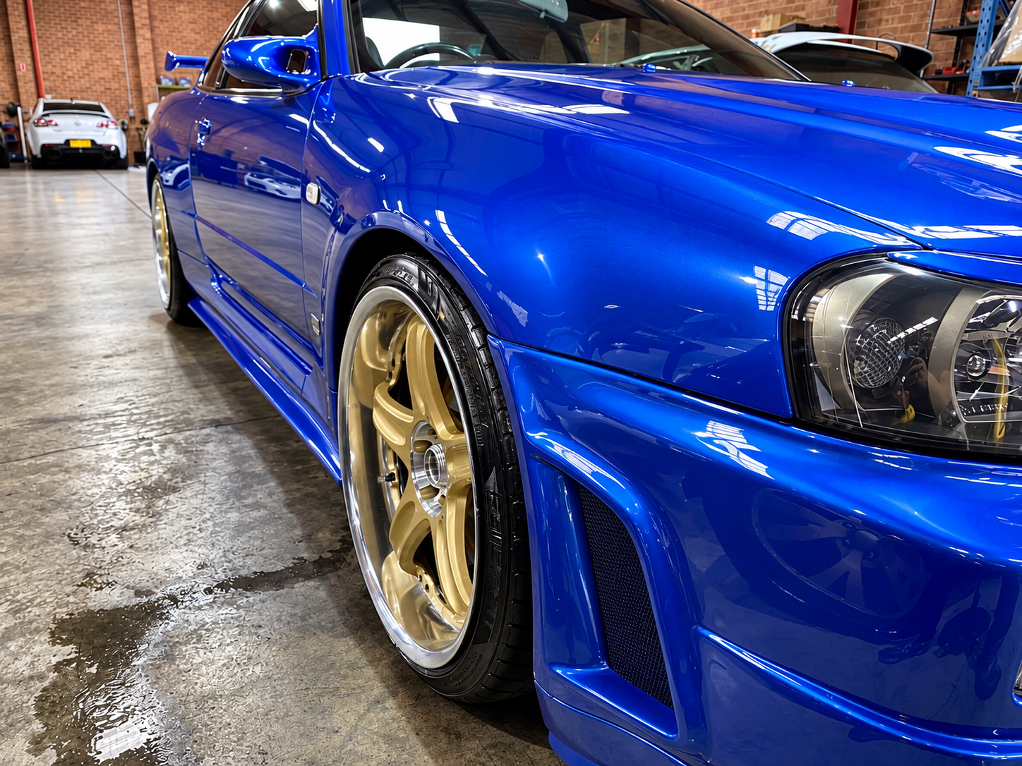 Close-up of a shiny blue sports car with gold rims, parked indoors in a garage with other cars and brick walls visible in the background.