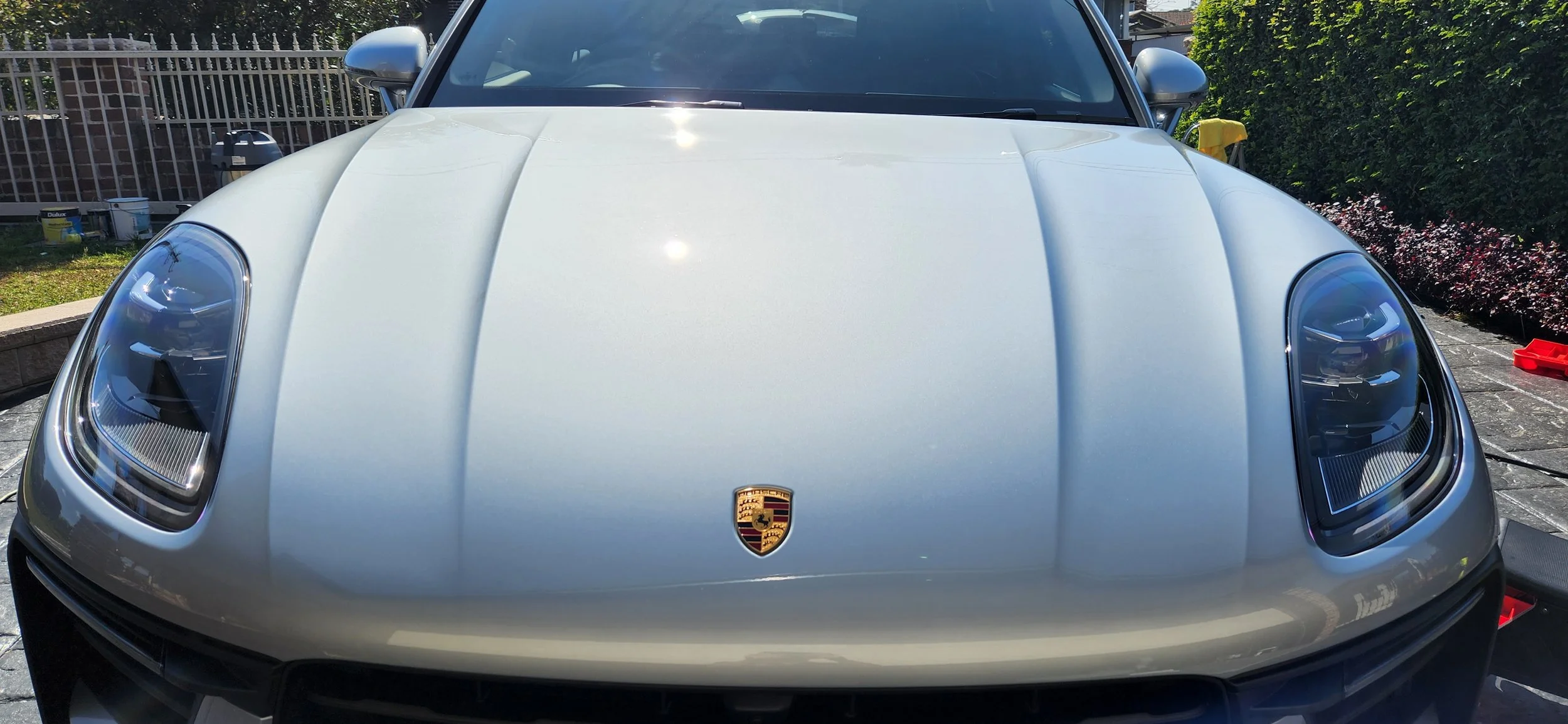 Front view of a silver Porsche sports car parked outdoors on paved ground, with a hedge and fence in the background.