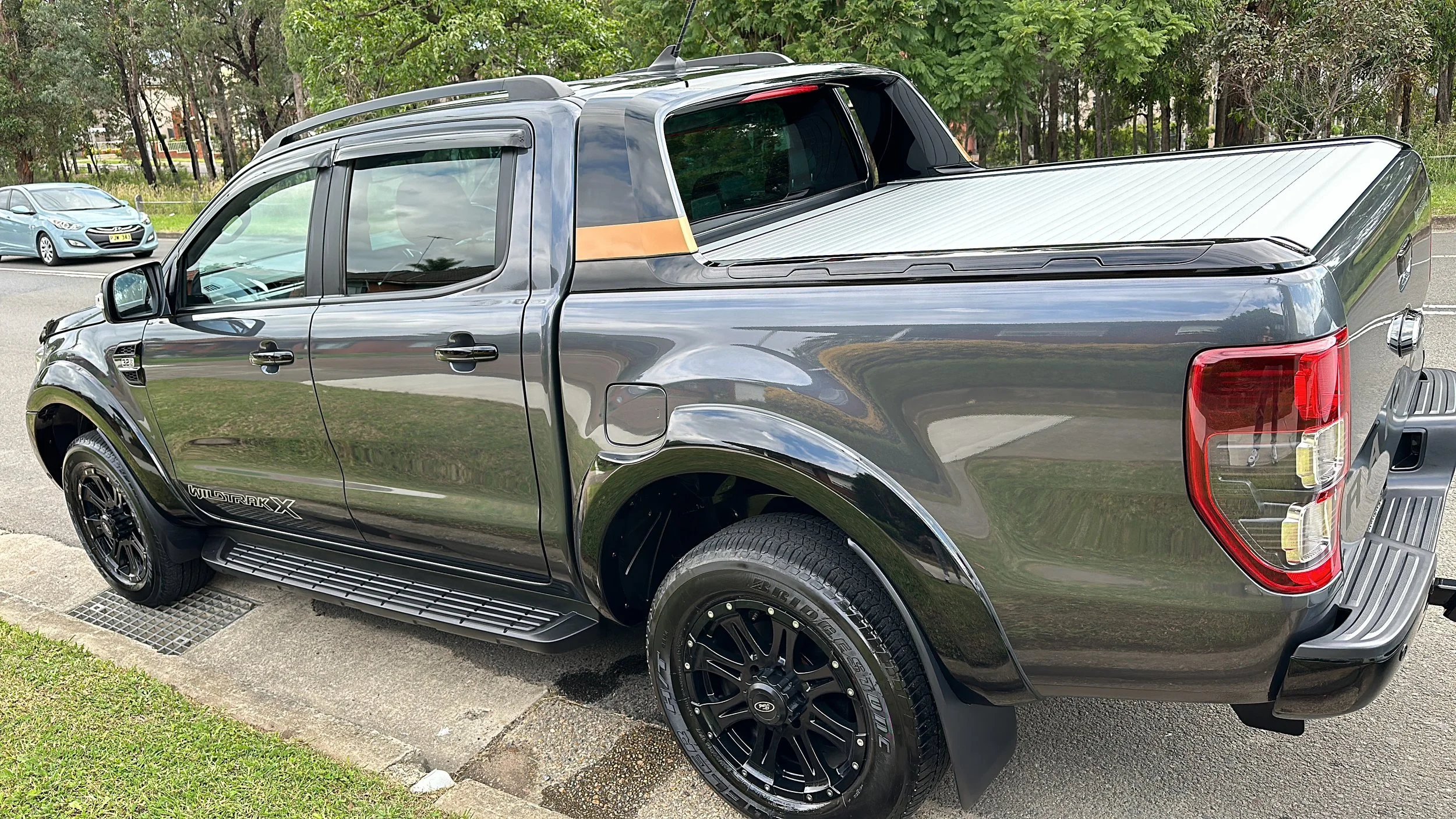 A black pickup truck parked on the side of a street with green trees in the background. The truck has black rims and a decorative cover on the bed.