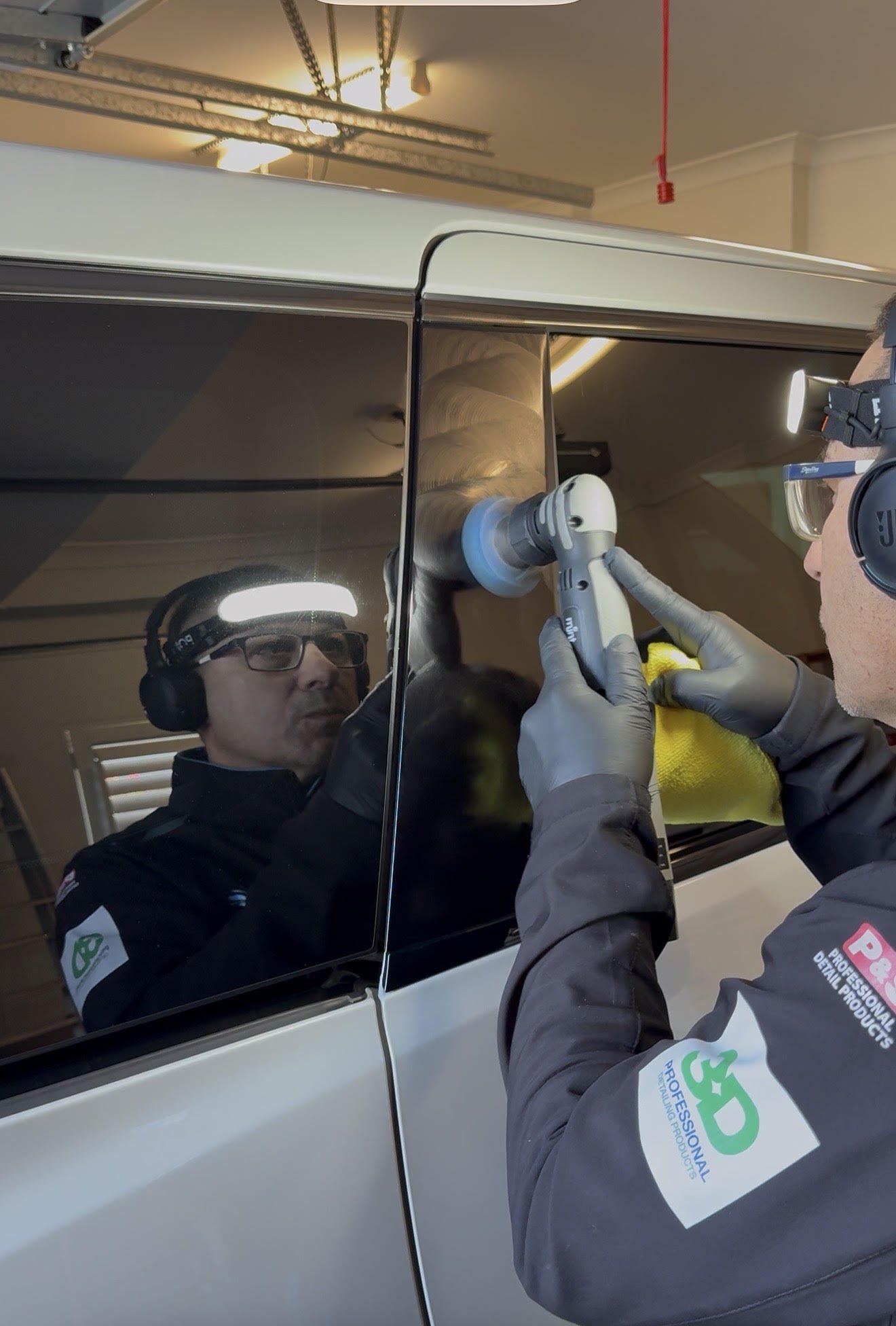 Peter A professional detailer wearing safety glasses, gloves, and a uniform with a logo, polishing a car's side mirror with a power buffer tool inside a garage.