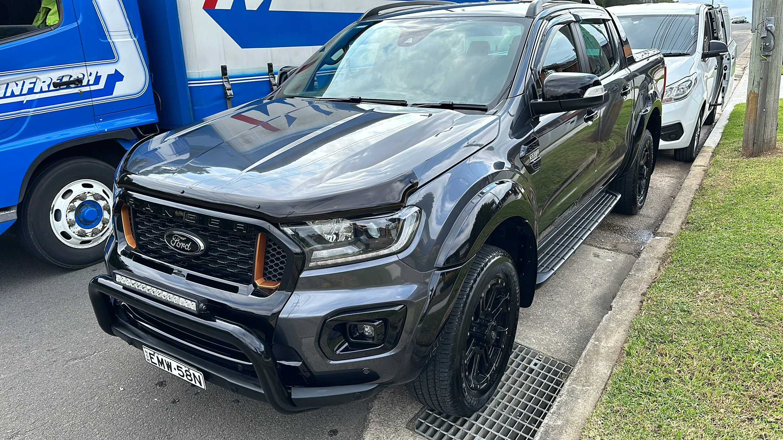 Black Ford Ranger pickup truck parked on the side of a street next to a white vehicle and a blue utility truck, with a grassy area and utility pole nearby.