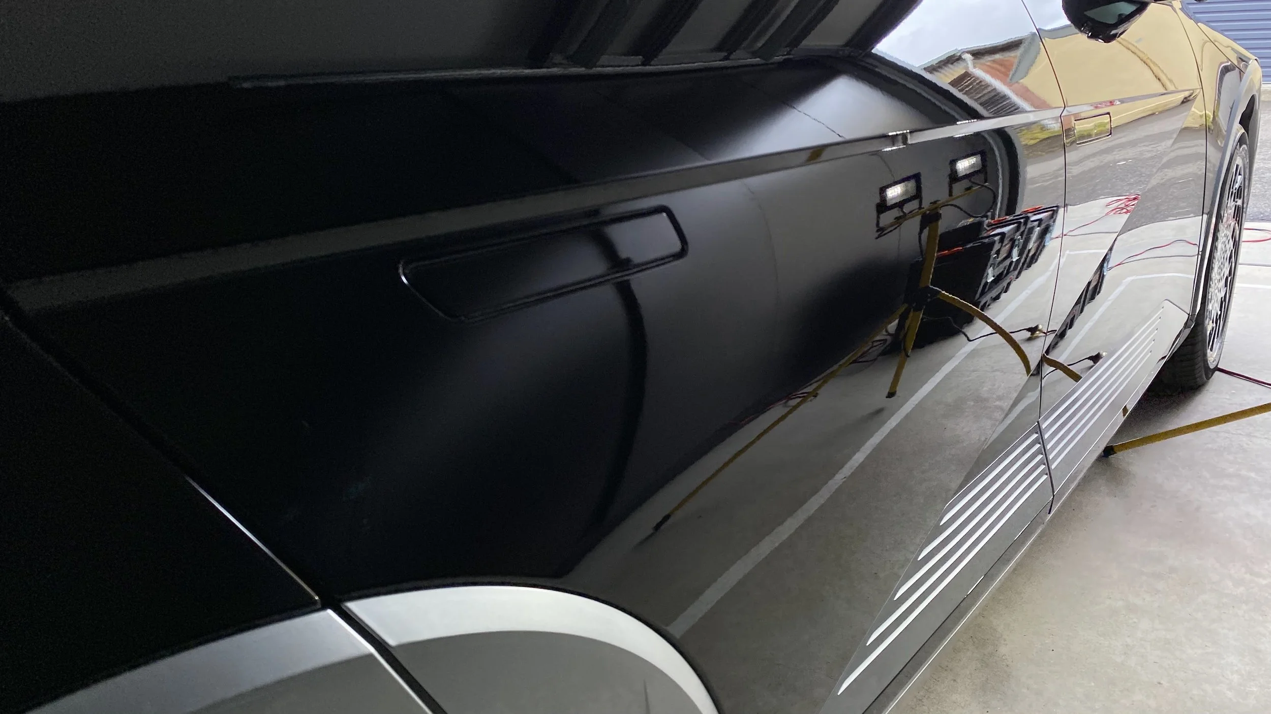 A black pickup truck with its hood open, parked indoors next to a yellow extension cord, with some equipment connected inside the truck's bed.