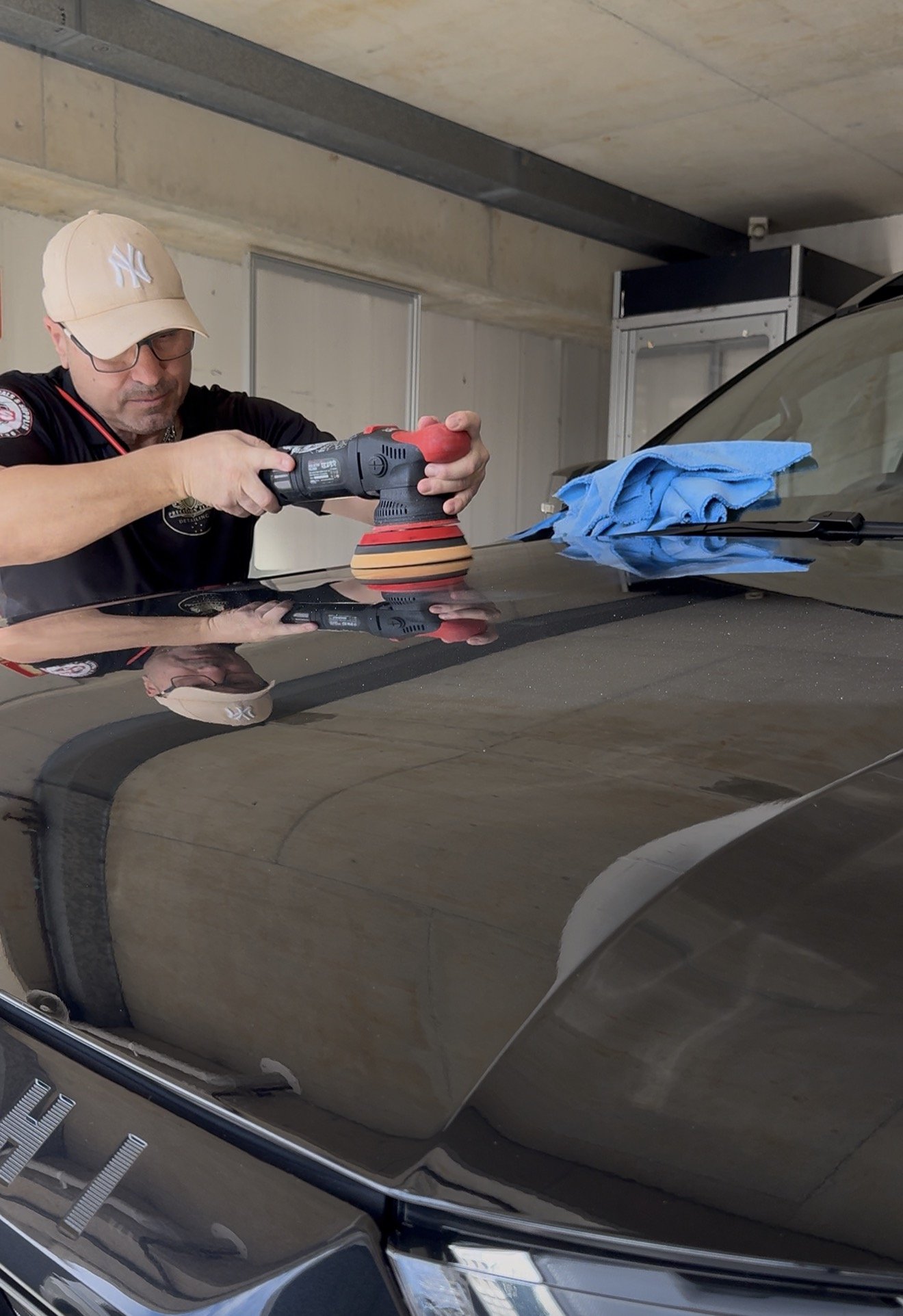 A person wearing a beige New York Yankees cap and glasses polishes the hood of a black car with a polishing machine. A blue cloth rests on the car's hood, and the car's reflection shows part of the person's face and the surrounding garage.