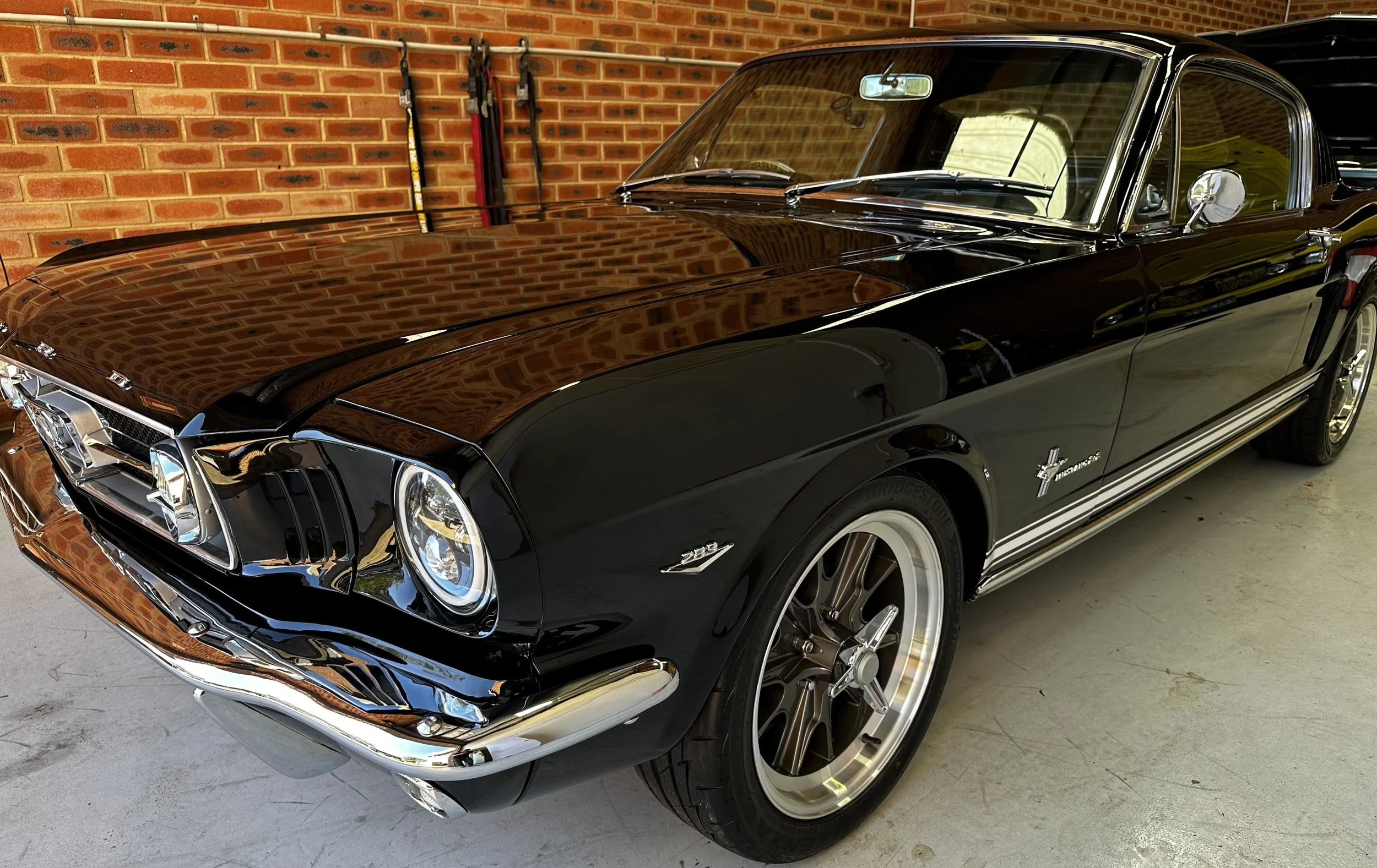 Black vintage Ford Mustang with chrome accents parked in a garage with a brick wall. The car's roof is black, and the body is shiny, reflecting the garage interior.